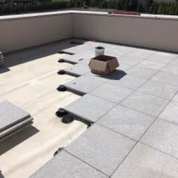 Montaż szarych płyt tarasowych na regulowanych wspornikach na dachu budynku, widoczny karton i wiadro z narzędziami.