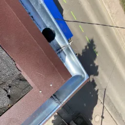 Widok z góry na narożnik nowo zainstalowanej, błyszczącej, metalowej rynny z odpływem, przylegającej do brązowej blachy na dachu pokrytym papą, z widokiem na ulicę z zielonymi oznaczeniami.