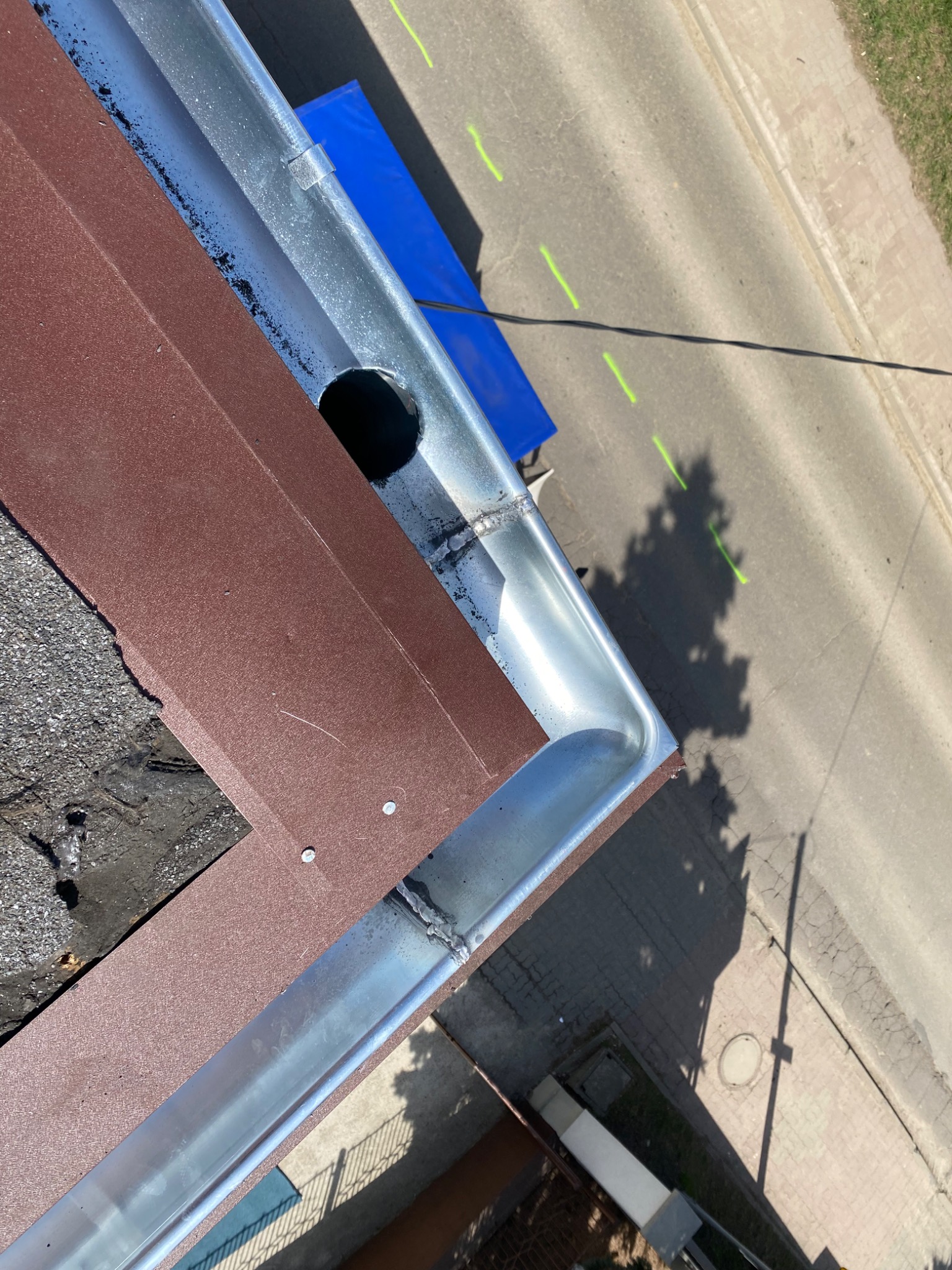 Widok z góry na narożnik nowo zainstalowanej, błyszczącej, metalowej rynny z odpływem, przylegającej do brązowej blachy na dachu pokrytym papą, z widokiem na ulicę z zielonymi oznaczeniami.