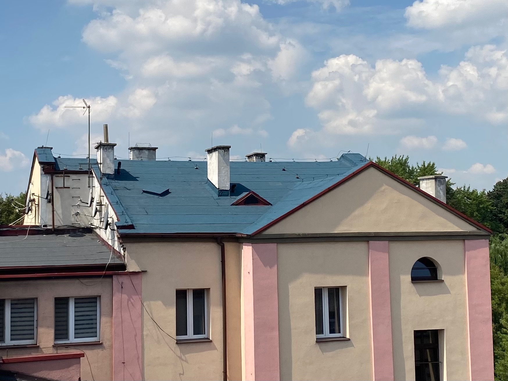 Niebieski dach budynku wielorodzinnego z białymi kominami na tle błękitnego nieba z chmurami. Widoczne anteny i instalacje na dachu sąsiedniego budynku.