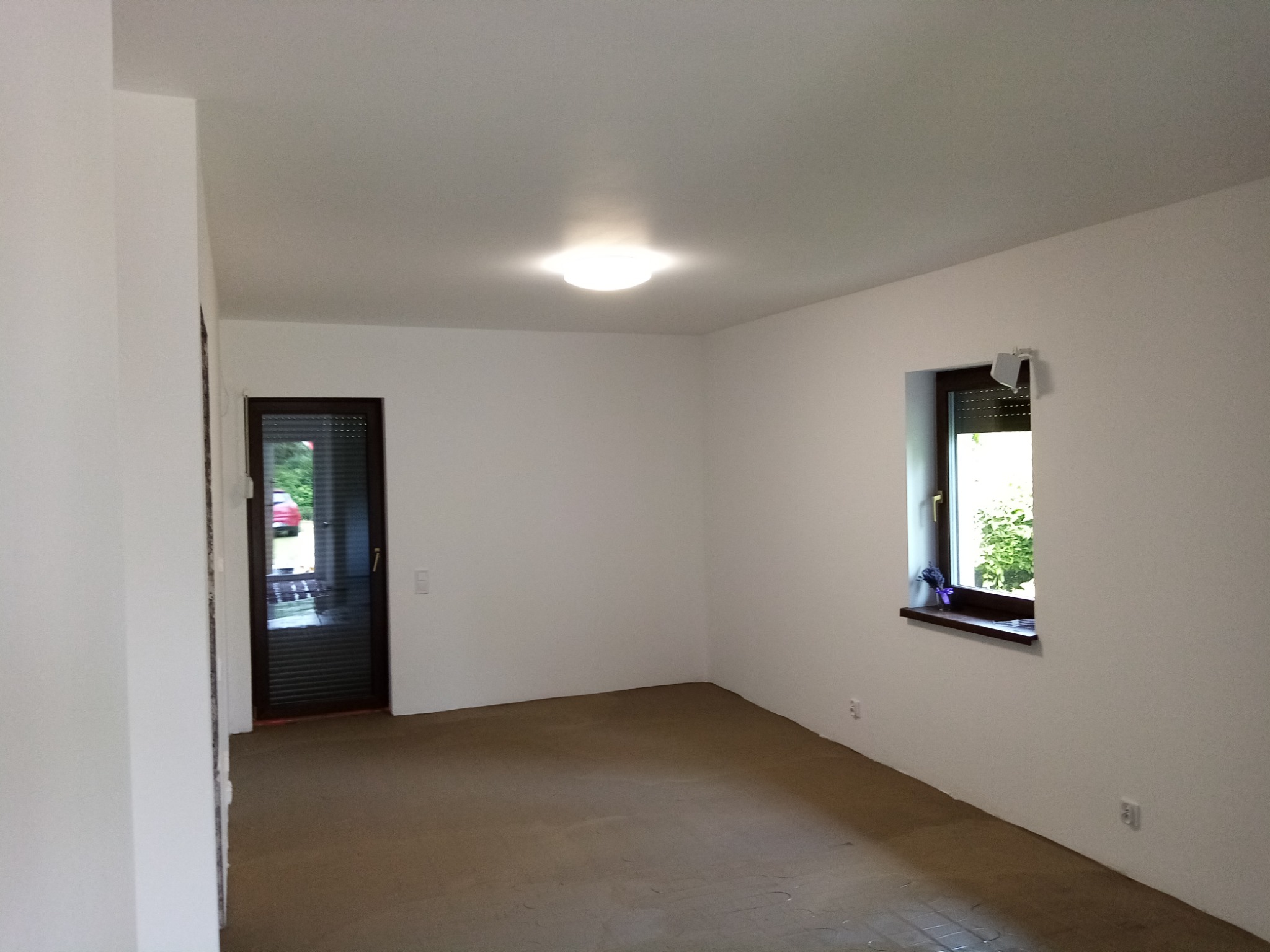 Pomalowane na biało wnętrze pokoju z widokiem na zewnątrz przez okno z roletami i czarną ramą, z podłogą przygotowaną pod dalsze prace wykończeniowe. Na parapecie stoi mały bukiet fioletowych kwiatów.