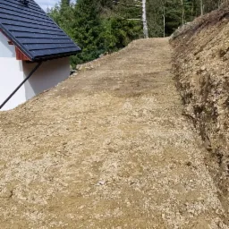 Wyrównany teren pod budowę domu jednorodzinnego z widocznym fragmentem dachu i drzewami w tle.