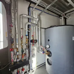 Węzeł cieplny z zbiornikiem akumulacyjnym, rurami w izolacji aluminiowej, zaworami i osprzętem, widoczne liczniki i okablowanie.