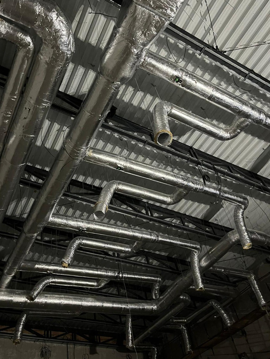 Srebrne, zaizolowane rury wentylacyjne zamontowane pod metalowym sufitem, widoczne czarne elementy konstrukcyjne i zwisające kable.