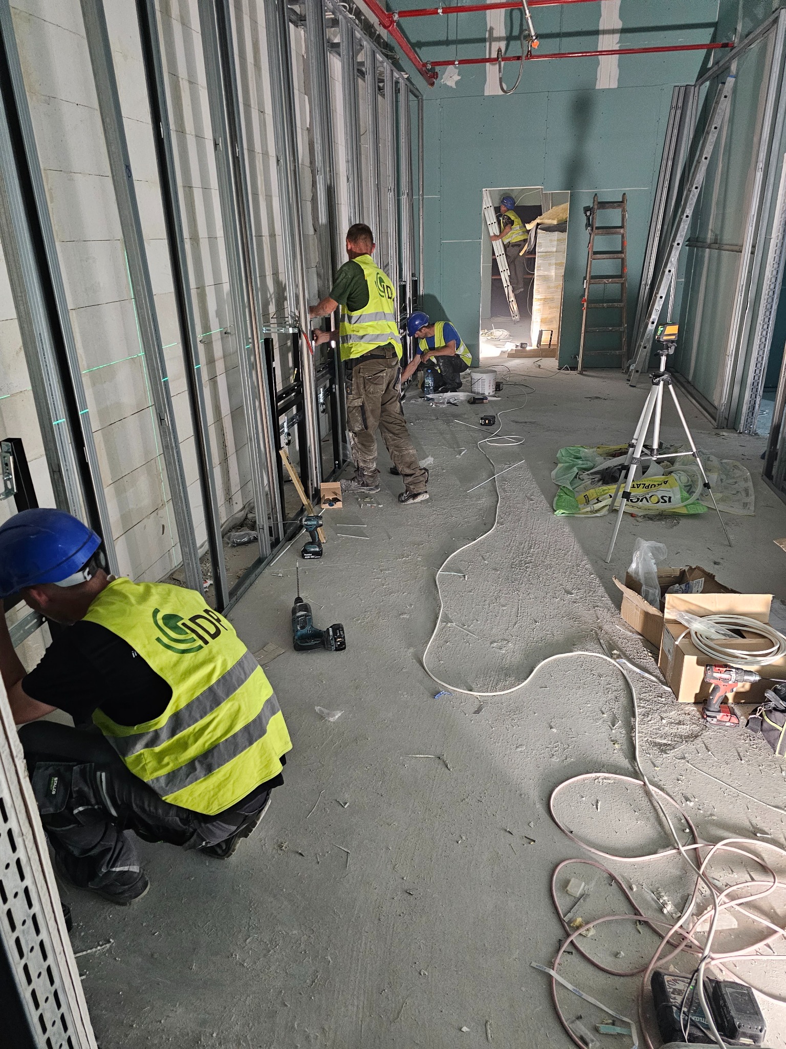 Prace wykończeniowe w Wilkowicach: montaż profili stalowych ścian działowych z instalacją elektryczną, widoczni pracownicy w odblaskowych kamizelkach i kaskach, narzędzia pracy oraz poziomica...