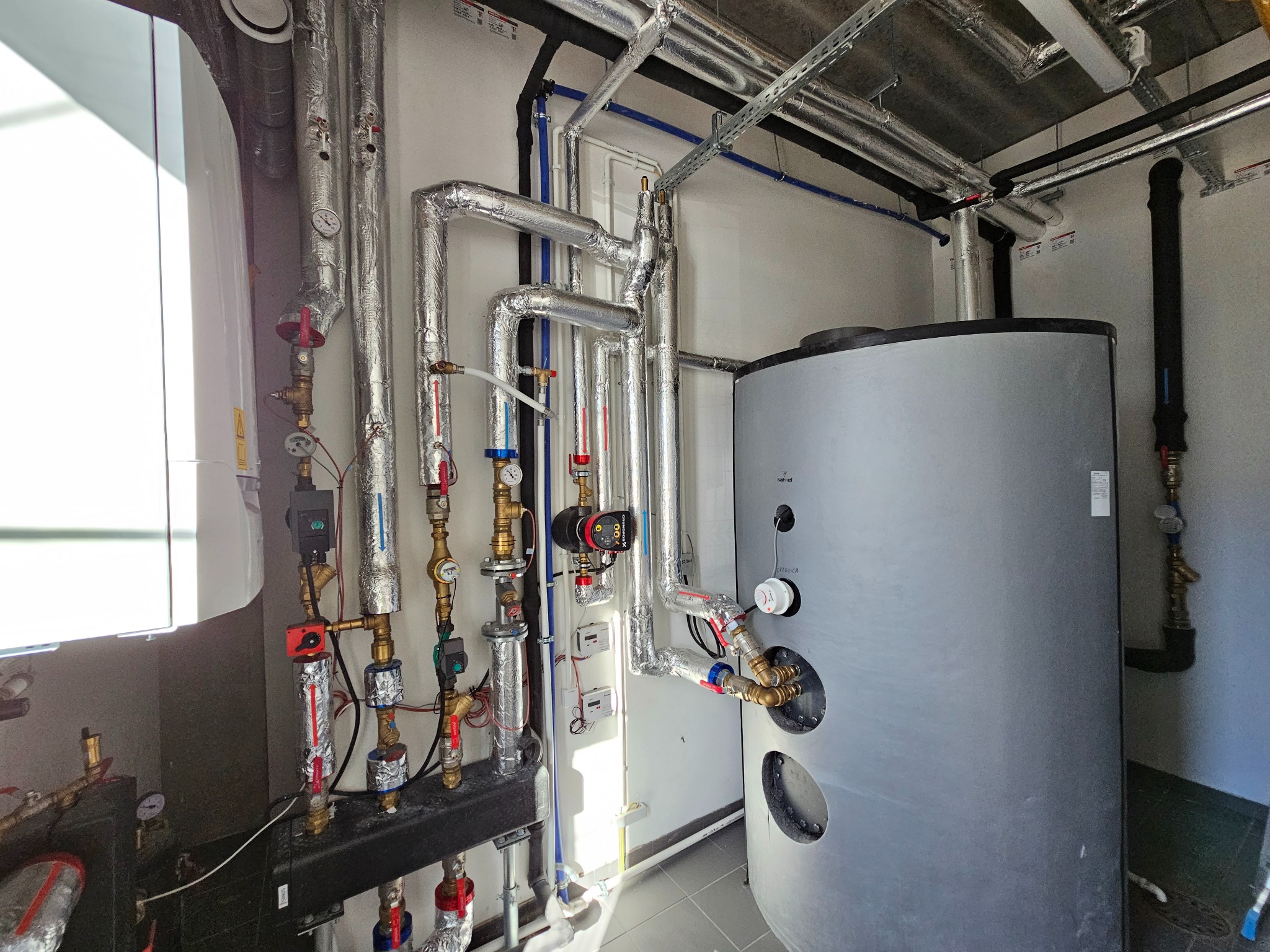 Węzeł cieplny z zbiornikiem akumulacyjnym, rurami w izolacji aluminiowej, zaworami i osprzętem, widoczne liczniki i okablowanie.
