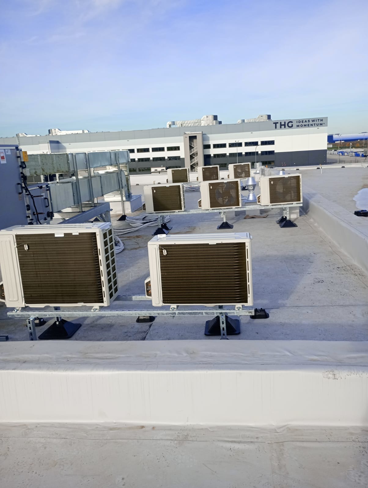 Rząd klimatyzatorów zamontowanych na dachu budynku przemysłowego z widocznym logo THG w tle.