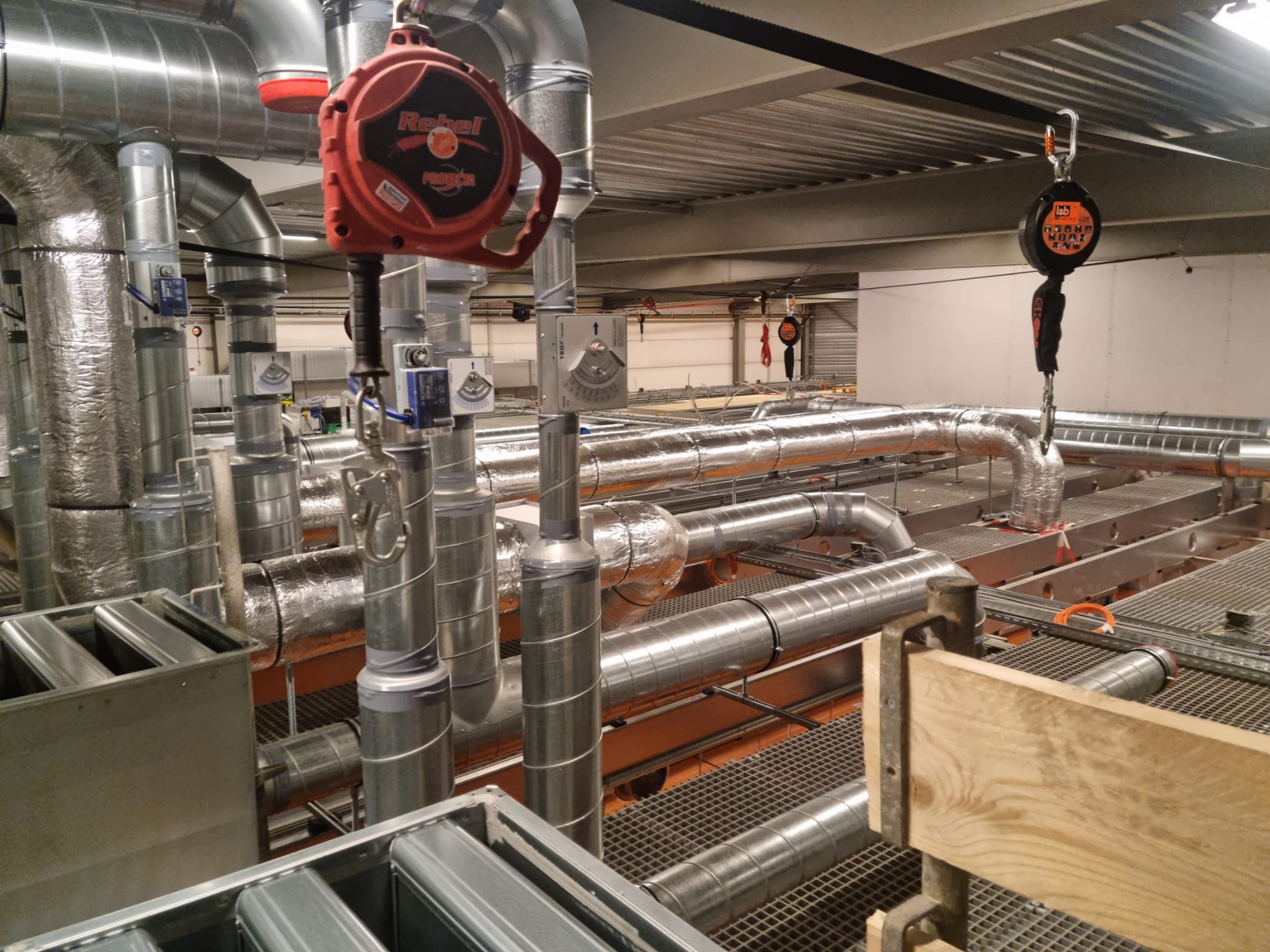 Skomplikowana instalacja rur wentylacyjnych i klimatyzacyjnych w przestrzeni technicznej pod sufitem, z widocznymi elementami bezpieczeństwa pracy na wysokości, takimi jak linki asekuracyjne...