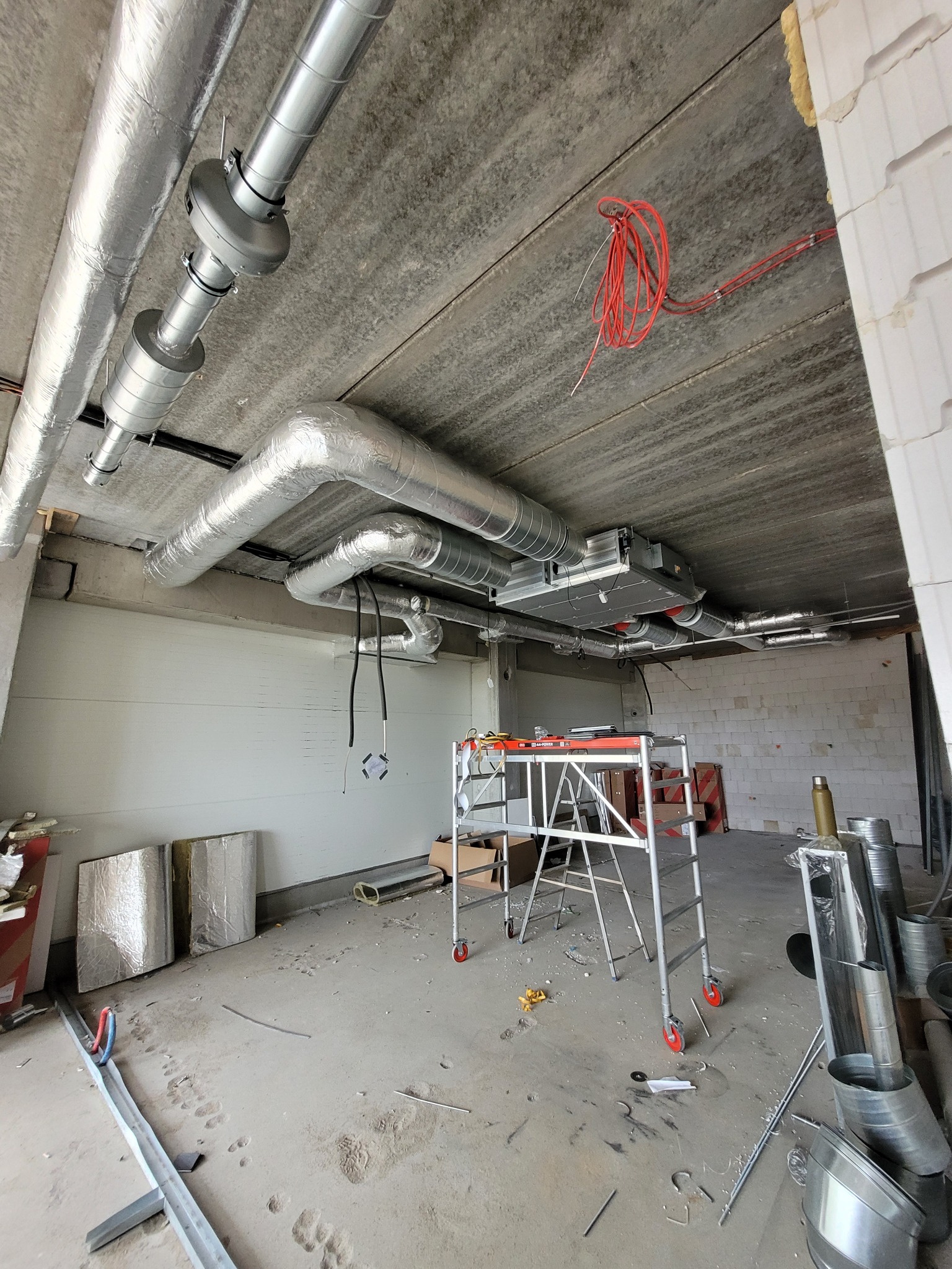 Wnętrze w trakcie montażu instalacji wentylacyjnej z widocznymi izolowanymi przewodami wentylacyjnymi pod sufitem, centralą wentylacyjną, rusztowaniem i narzędziami na podłodze.