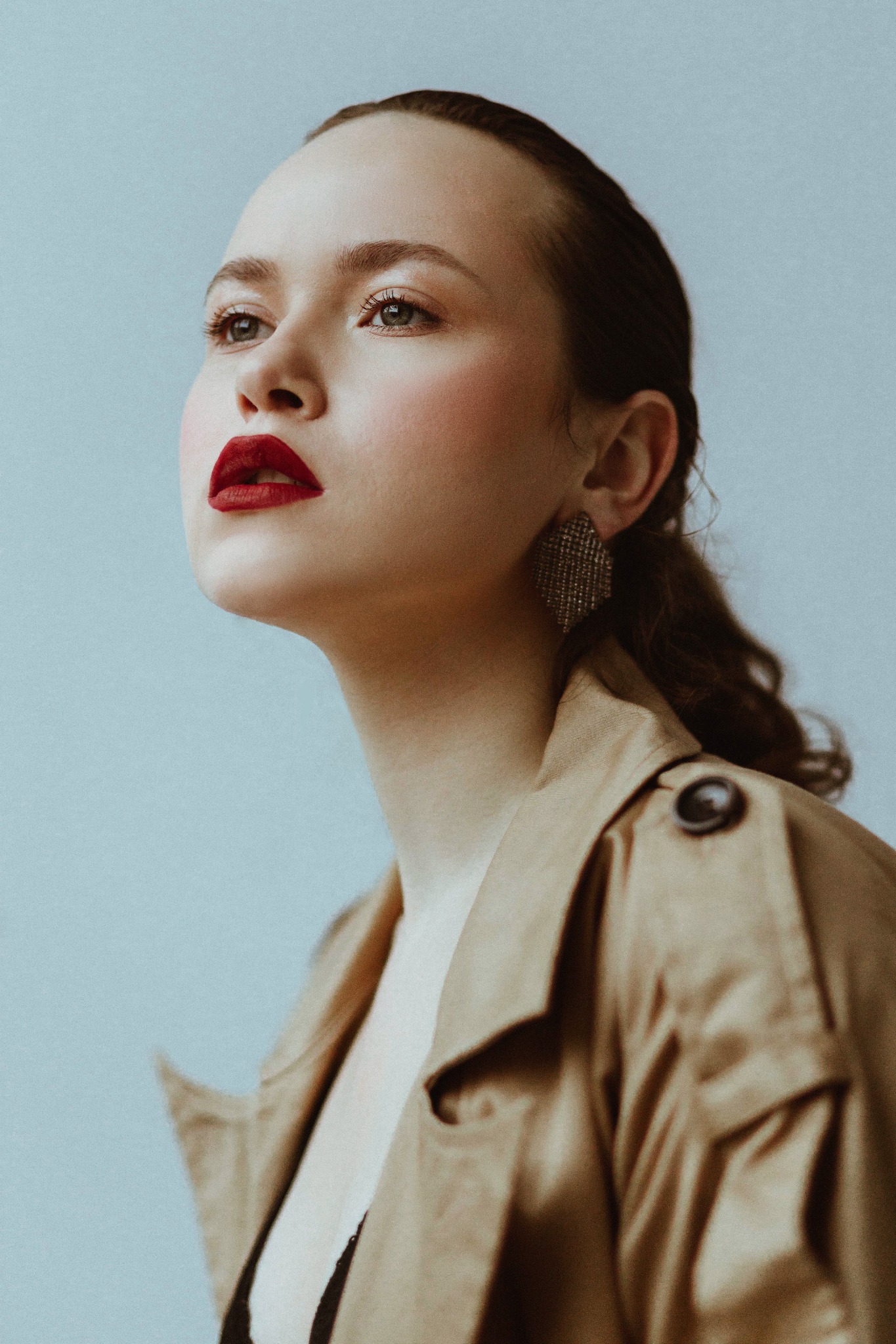 Portret kobiety z mocnym makijażem ust, w beżowym płaszczu, z dużym kolczykiem w uchu, spoglądającej w górę na jasnym tle.