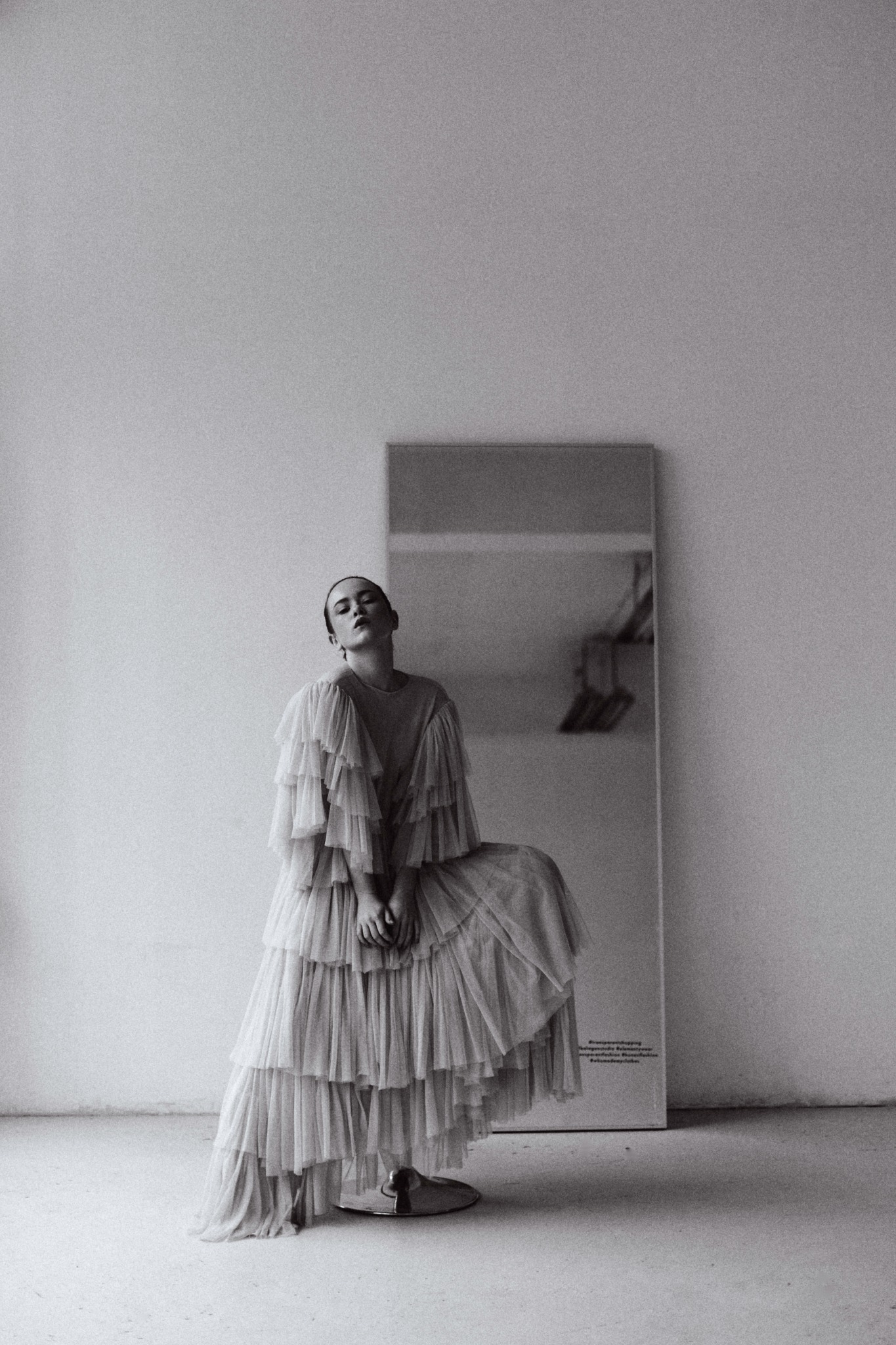 Czarno-białe zdjęcie kobiety w długiej, warstwowej sukni z falbanami, pozującej w minimalistycznym wnętrzu z lustrem w tle.