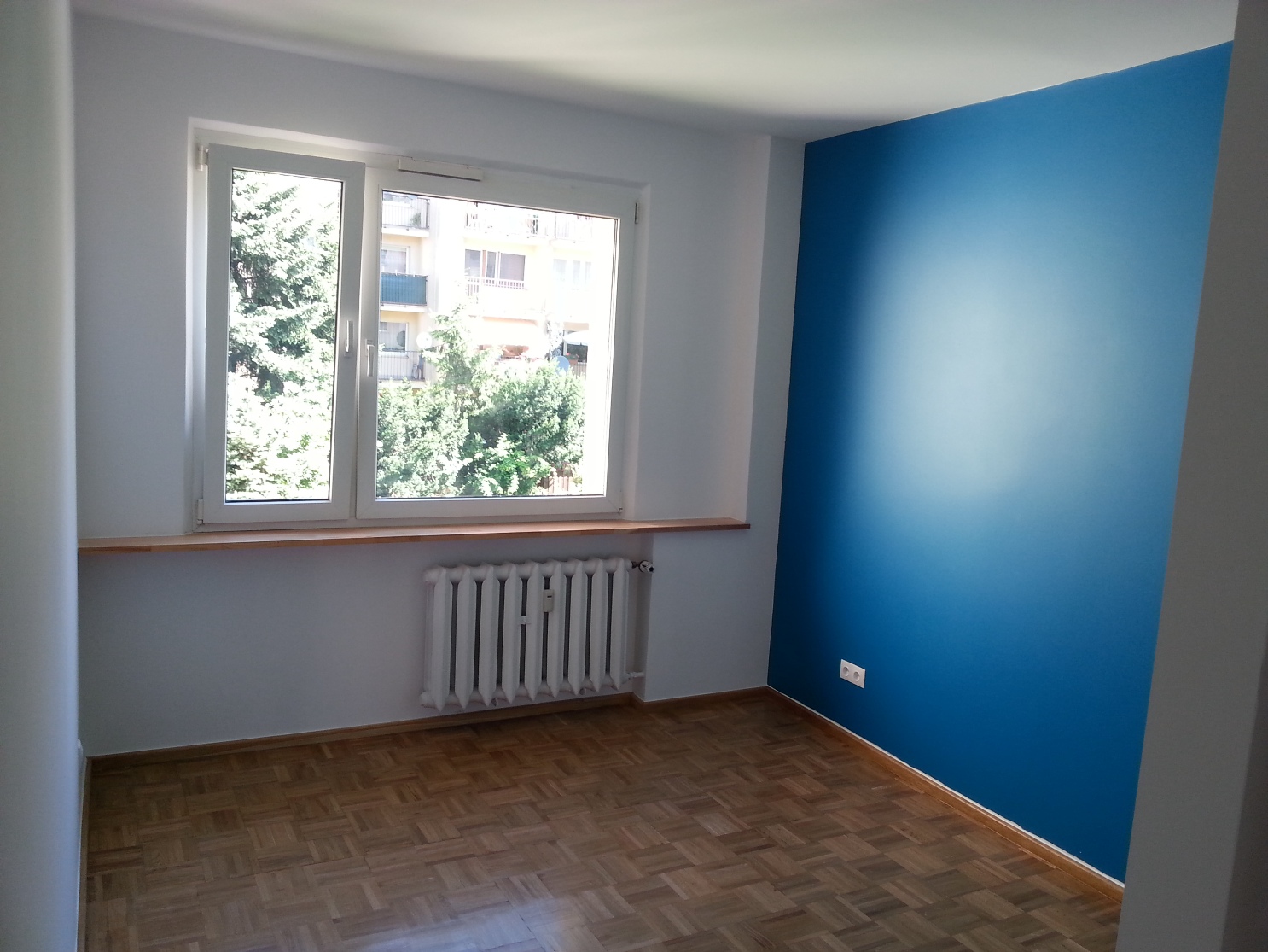 Pomalowane na biało wnętrze pokoju z jedną ścianą w odcieniach niebieskiego, drewnianą podłogą parkietową i białym grzejnikiem pod oknem.