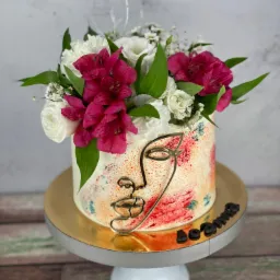 Tort zdobiony abstrakcyjnym motywem twarzy z metalu i bukietem z białych i różowych kwiatów, umieszczony na białym stojaku z napisem 'Joanna'.