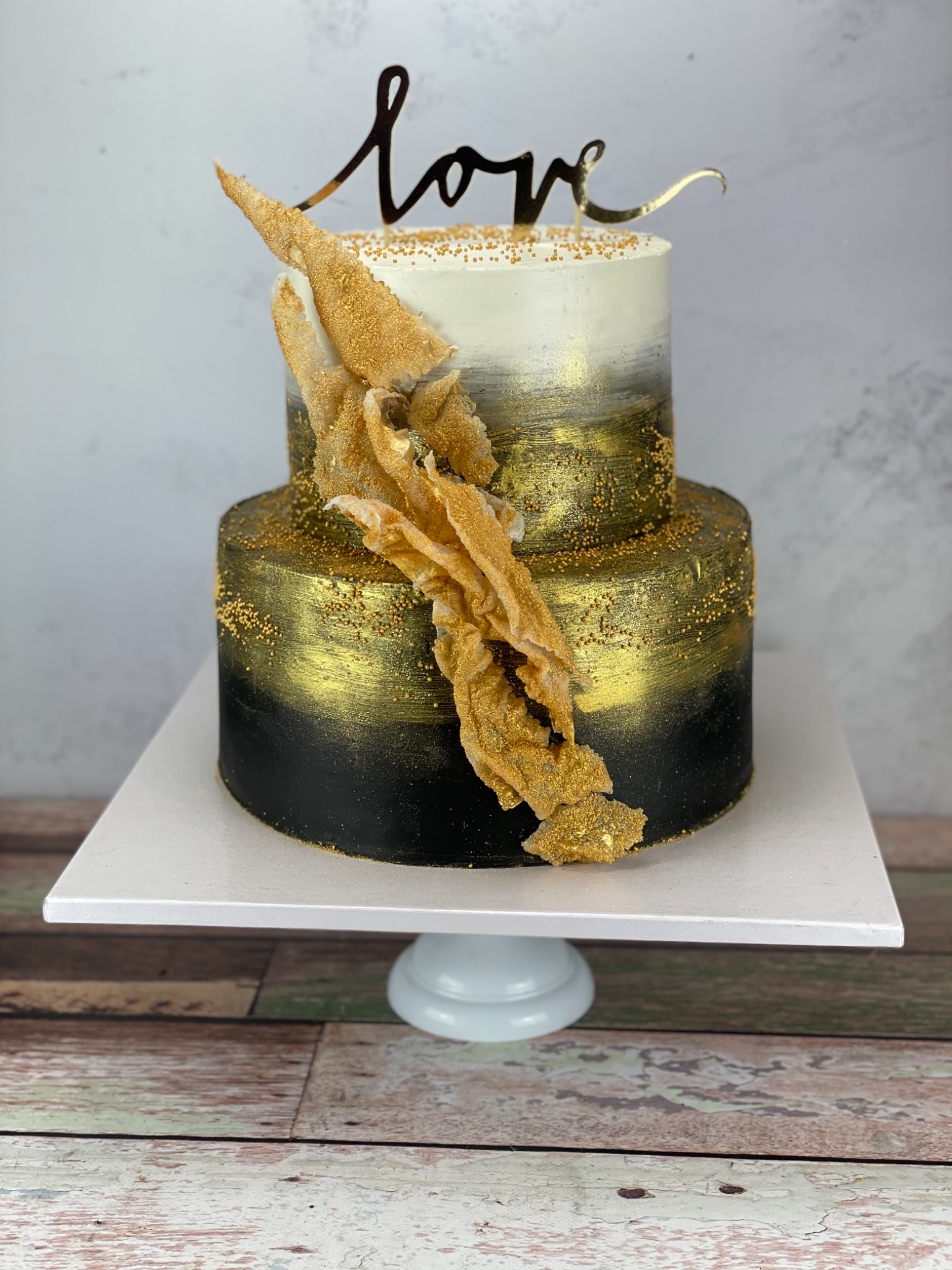 Dwupoziomowy tort weselny w kolorach czerni i złota, ozdobiony abstrakcyjną dekoracją z jadalnego złota i napisem 'love'.