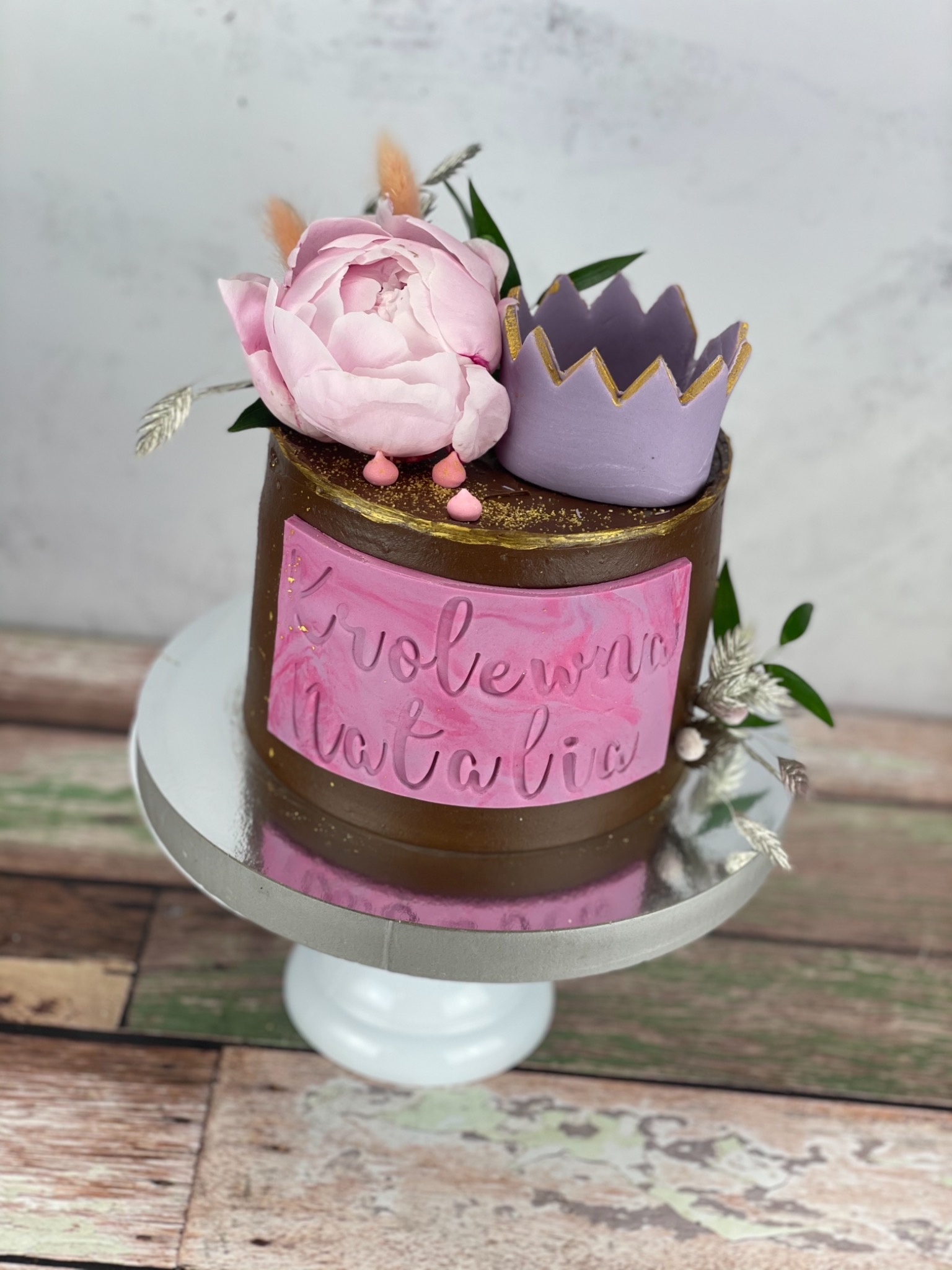 Tort dwuwarstwowy z czekoladowym kremem, ozdobiony różową piwonią, liliową koroną z brokatowymi krawędziami i tabliczką z marmurkowym wzorem oraz napisem 'Krolewna Natalia', umieszczony na srebrnej...