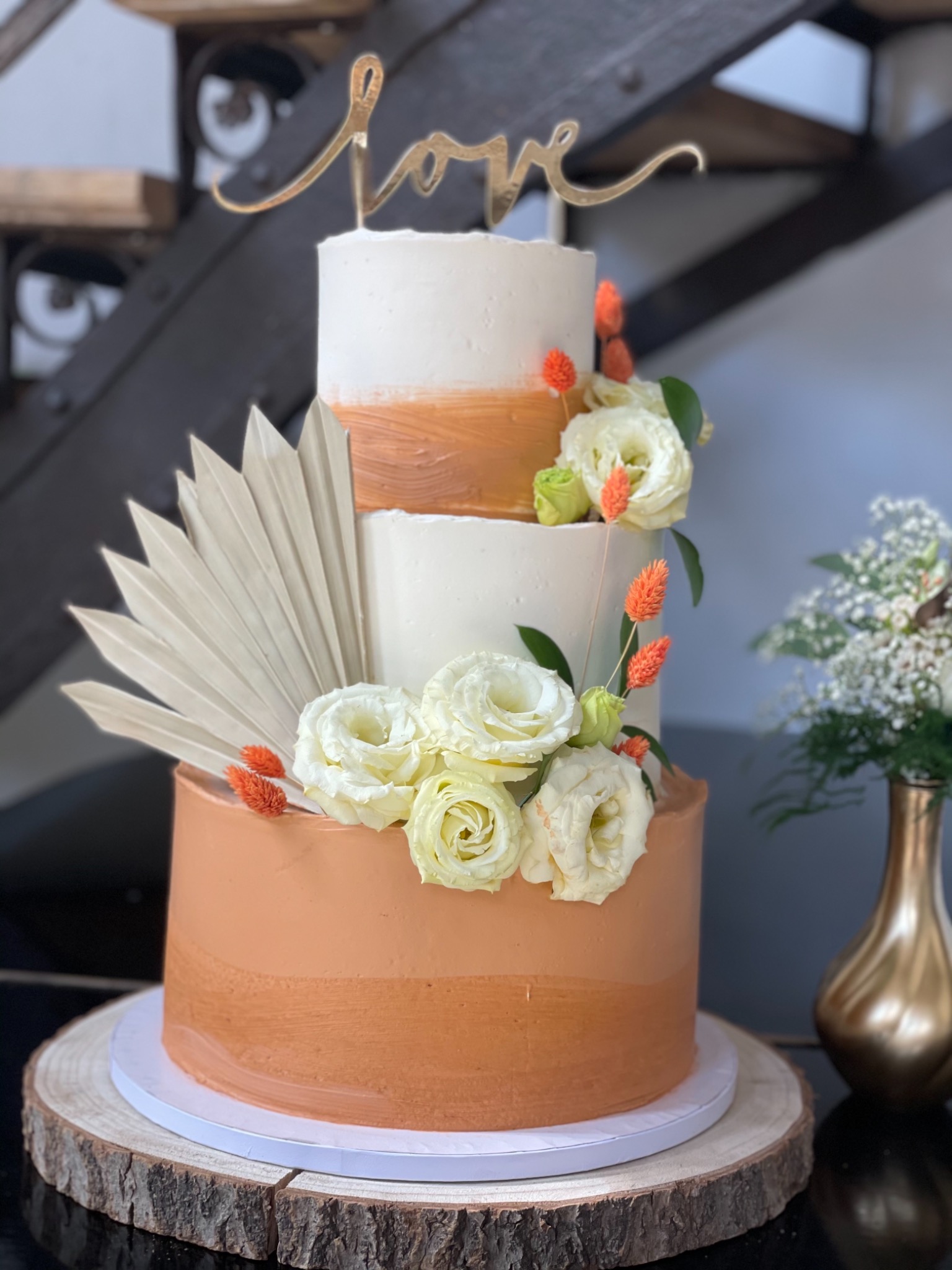 Trzypiętrowy tort weselny z motywem boho, zdobiony białymi różami, suszonymi pomarańczowymi kwiatami i wachlarzem z liści palmowych, umieszczony na drewnianym plastrze.