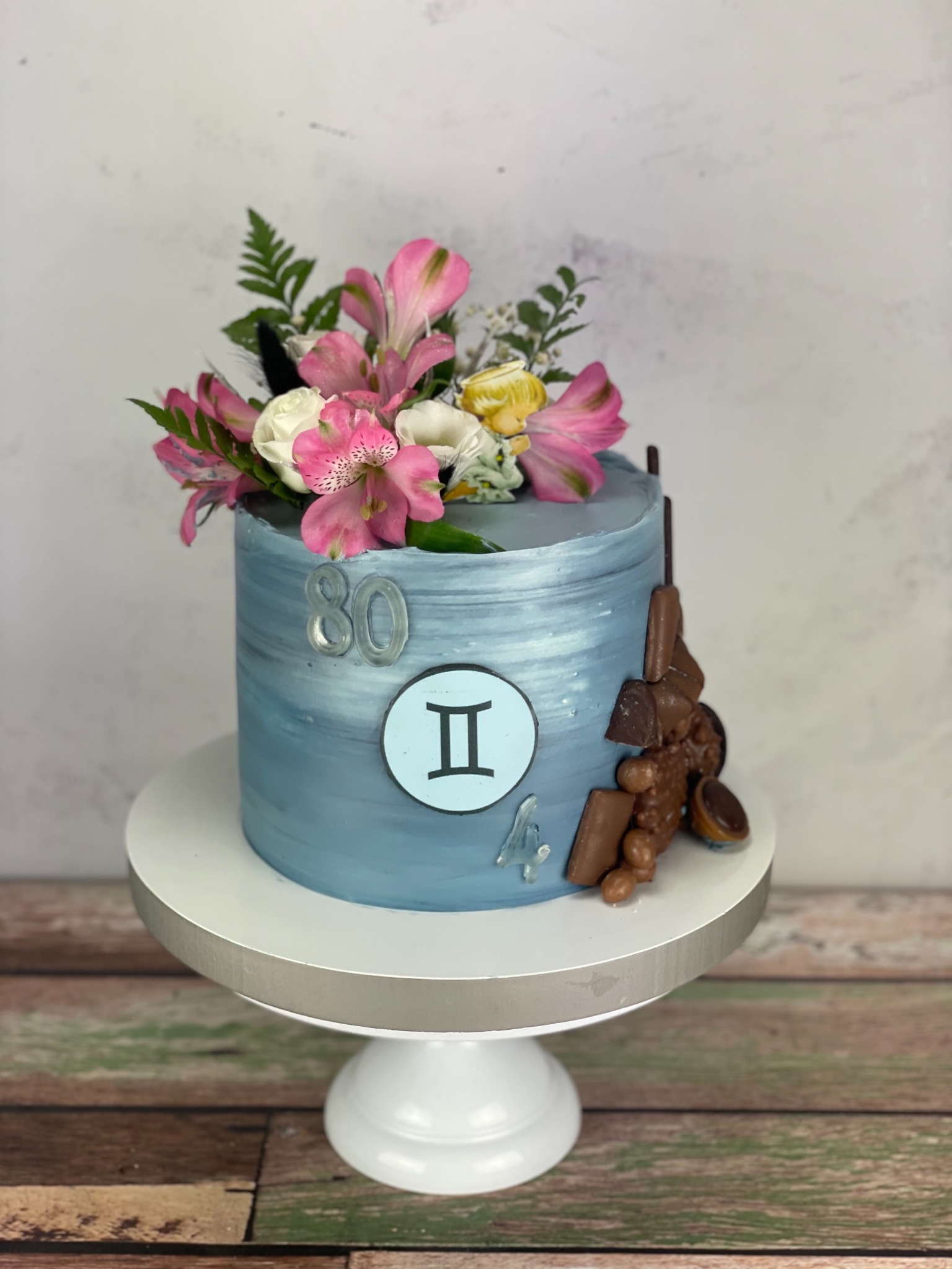 Tort urodzinowy w odcieniach błękitu, udekorowany świeżymi kwiatami, czekoladą i symbolem Bliźniąt, oznaczony cyfrą 80 i 4, na białym stojaku.