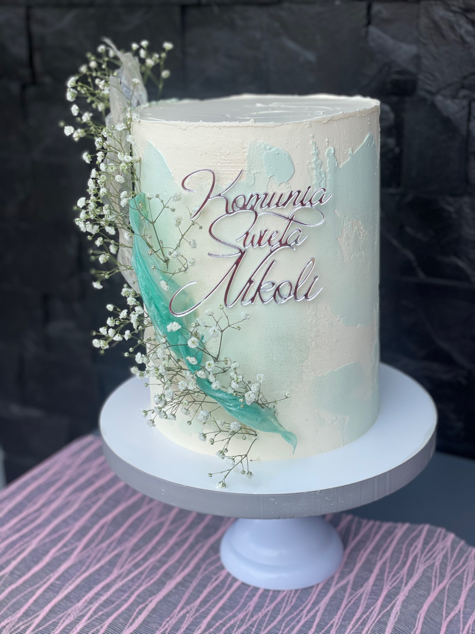 Tort komunijny w odcieniach bieli i błękitu, zdobiony gipsówką i srebrnym napisem 'Komunia Święta Nikoli', ustawiony na białej paterze.