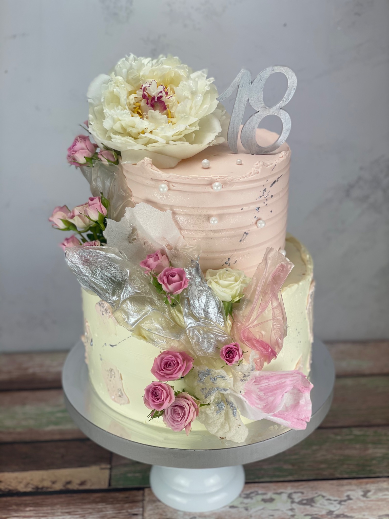Dwupoziomowy tort urodzinowy na 18-te urodziny, zdobiony kremem w odcieniach różu i żółci, świeżymi różami, dużą piwonią i srebrną dekoracją z numerem 18.