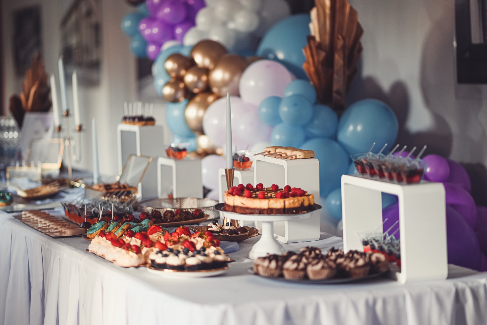 Stół z deserami na przyjęciu, udekorowany balonami w odcieniach fioletu, błękitu i złota; widoczne ciasta z owocami, babeczki i inne słodkości.