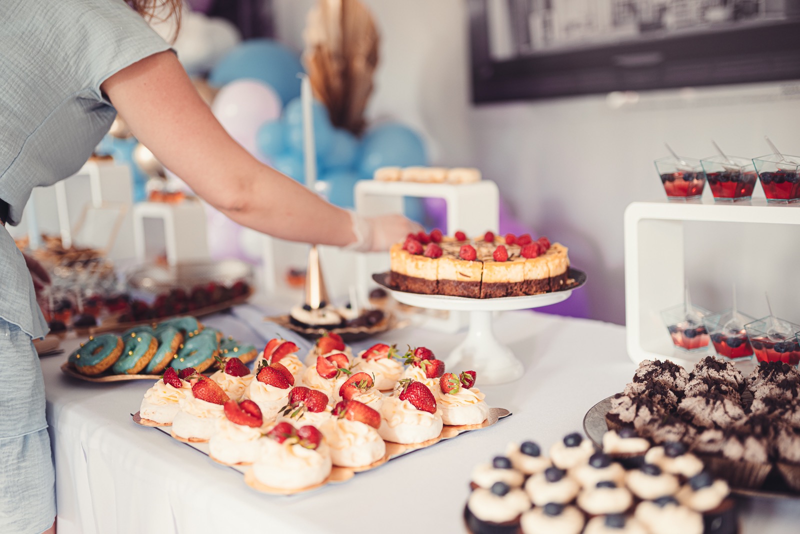 Stół z różnorodnymi deserami: mini bezy z truskawkami, turkusowe donuty z posypką, babeczki czekoladowe z borówkami, sernik z malinami na paterze i galaretki z owocami, przygotowywane przez osobę...
