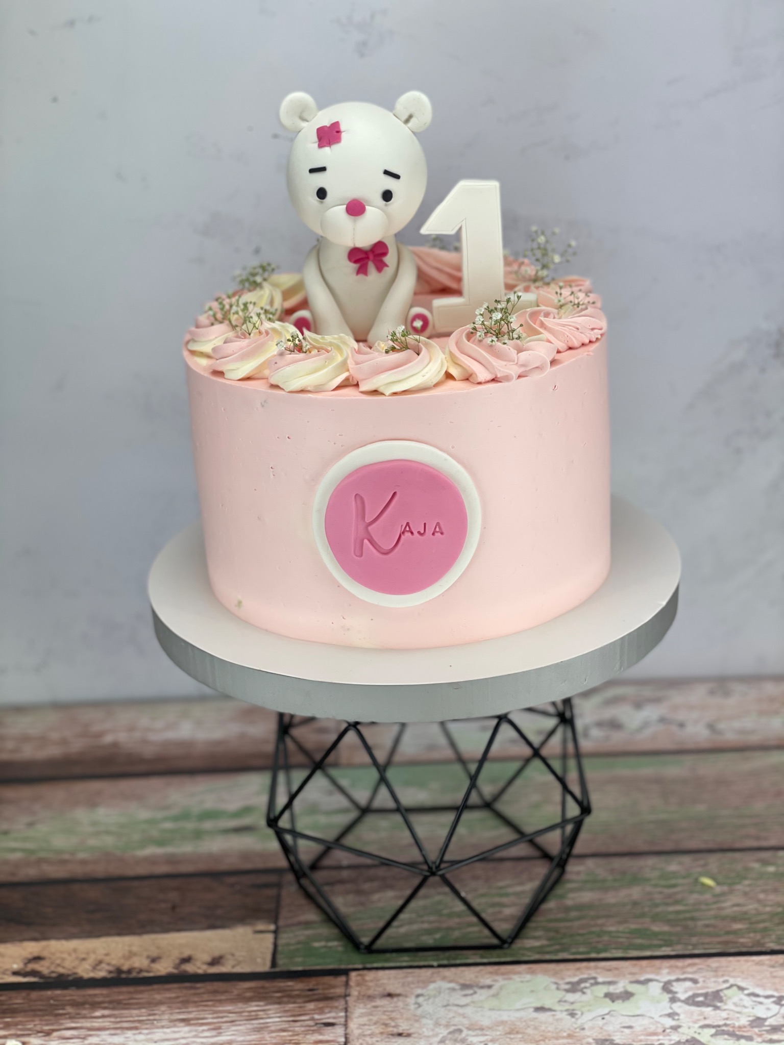 Różowy tort urodzinowy z białym misiem z lukru, cyfrą '1' i imieniem 'Kaja', udekorowany kremem i drobnymi kwiatkami, na metalowym stojaku.
