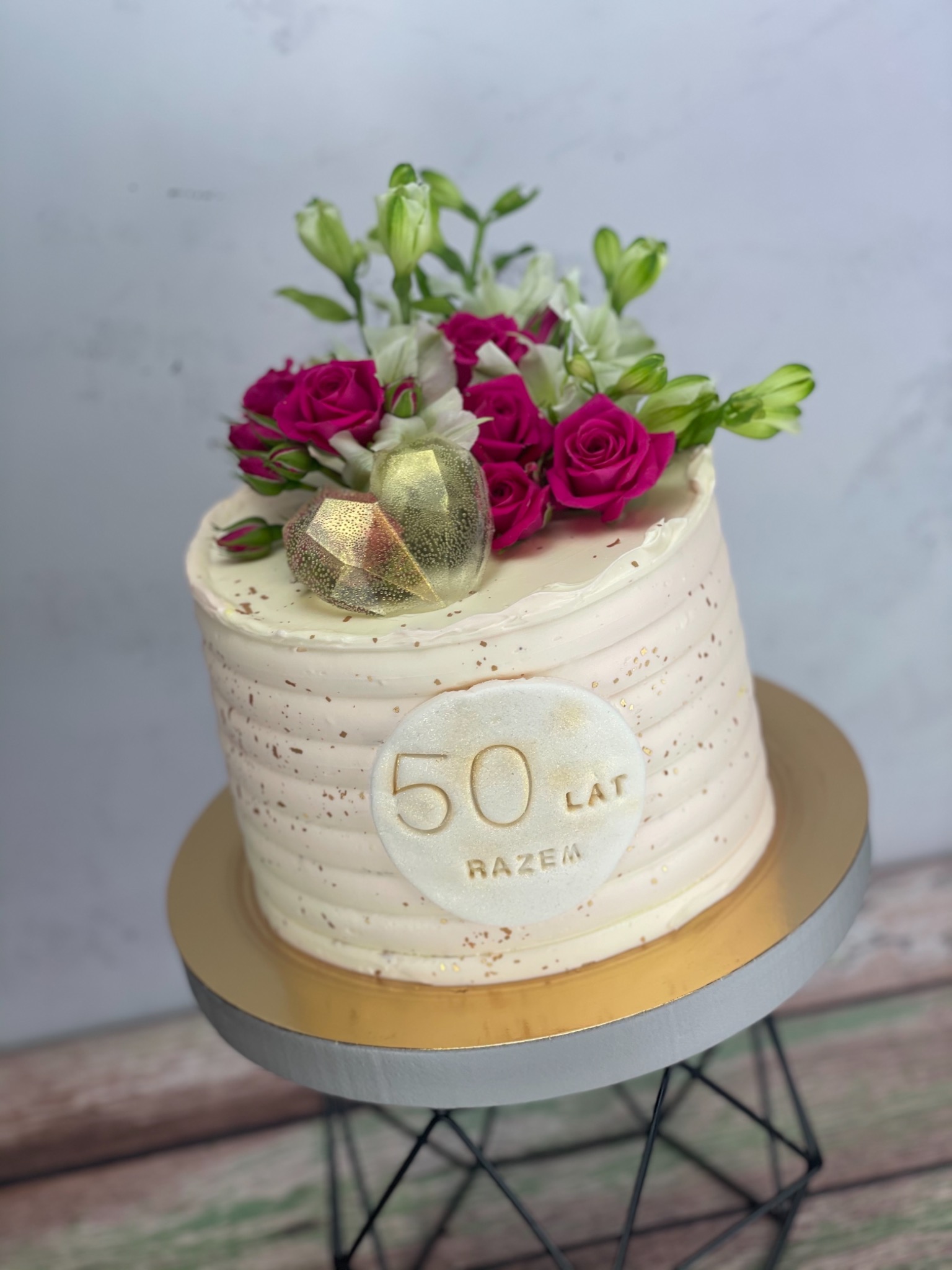 Tort na 50-lecie z białym kremem, złotymi akcentami, dekoracją z różowych róż i białych alstromerii oraz złotym napisem '50 lat razem', umieszczony na złotym podeście i czarnym metalowym stojaku.