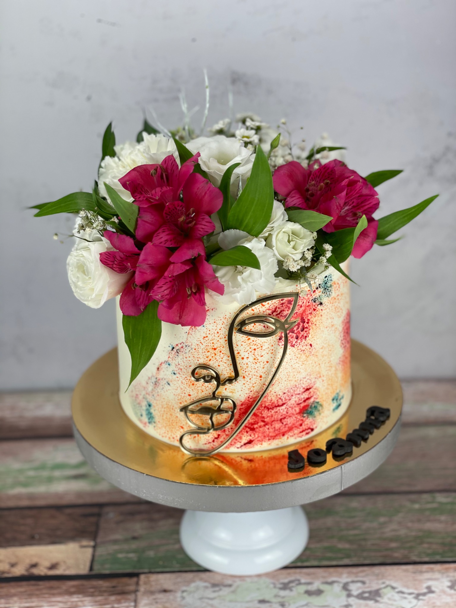 Tort zdobiony abstrakcyjnym motywem twarzy z metalu i bukietem z białych i różowych kwiatów, umieszczony na białym stojaku z napisem 'Joanna'.