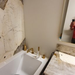 Instal-rem - Biała wanna z szarą miską wewnątrz, złota bateria i dozownik mydła na brzegu, obok umywalka z marmurowym blatem i złotymi uchwytami, ściana wyłożona marmurem.