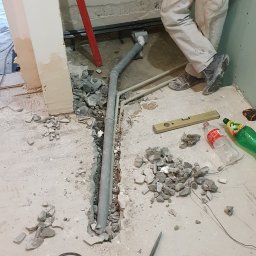 Instal-rem - Wykuta bruzda w podłodze z szarą rurą kanalizacyjną, gruz, poziomica, perforator Metabo, instalator w roboczych spodniach oparty o ścianę.