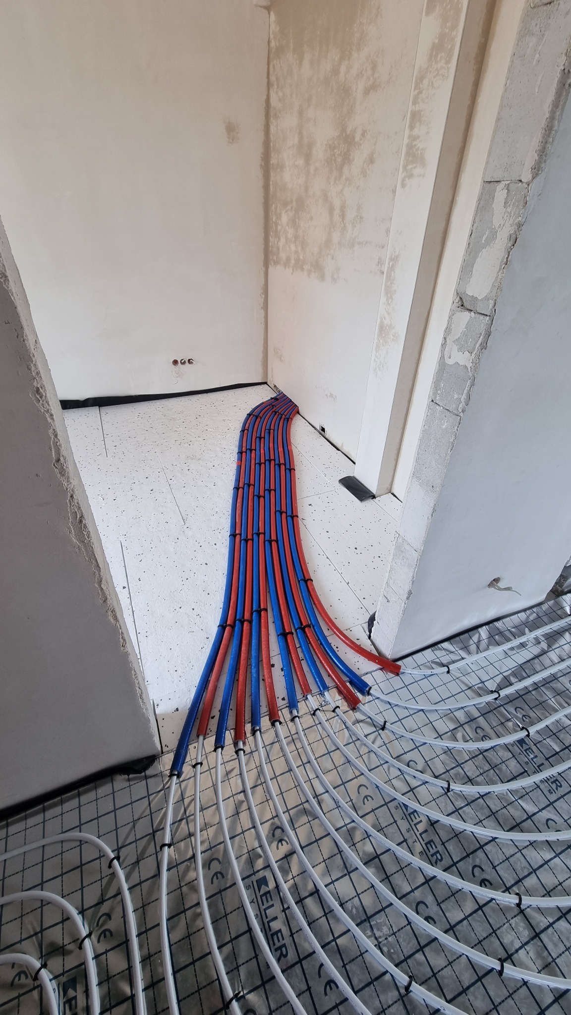 Rozłożone rury ogrzewania podłogowego na folii z widocznymi nieotynkowanymi ścianami w tle, z pionowym układem rur czerwonych i niebieskich doprowadzających.