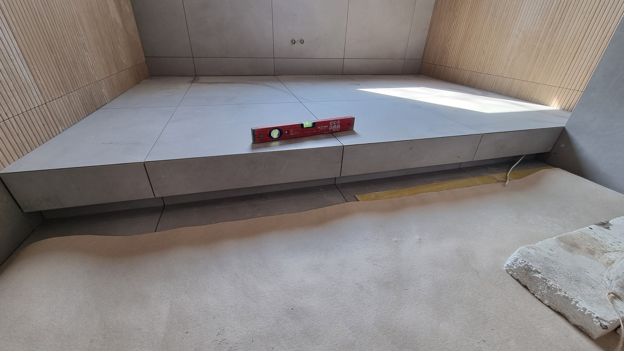 Fragment łazienki w trakcie wykańczania: szare płytki na podwyższeniu, poziomica na krawędzi, zabezpieczona podłoga, drewnopodobne panele na ścianie.