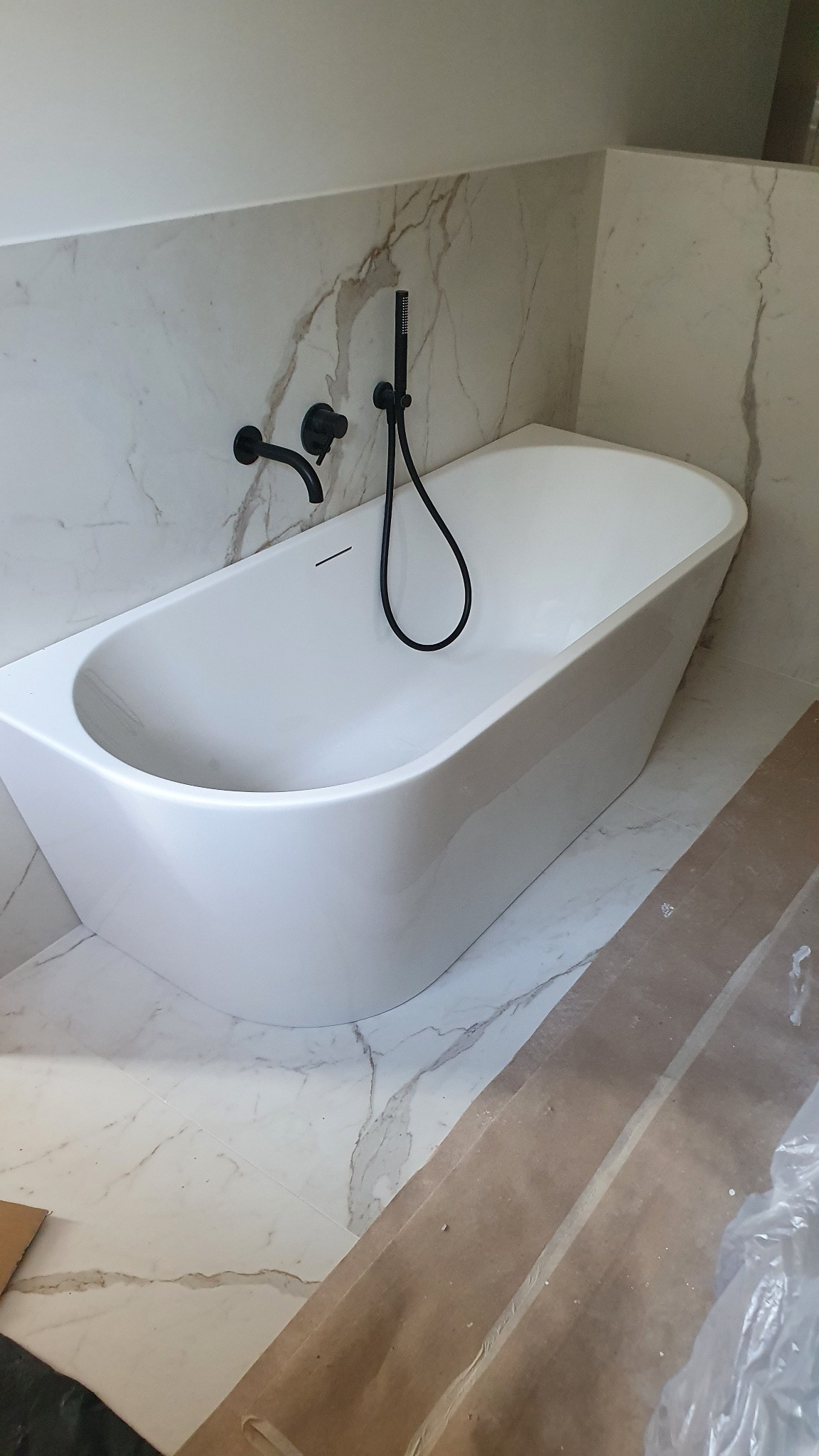 Nowoczesna, wolnostojąca wanna z czarną armaturą na tle marmurowej ściany, zabezpieczona podłoga podczas montażu.