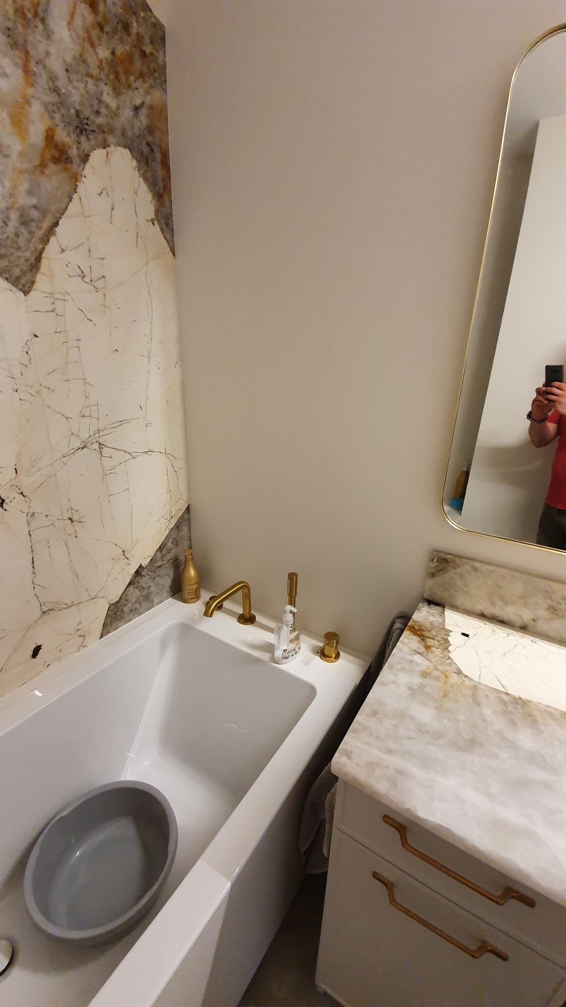 Biała wanna z szarą miską wewnątrz, złota bateria i dozownik mydła na brzegu, obok umywalka z marmurowym blatem i złotymi uchwytami, ściana wyłożona marmurem.