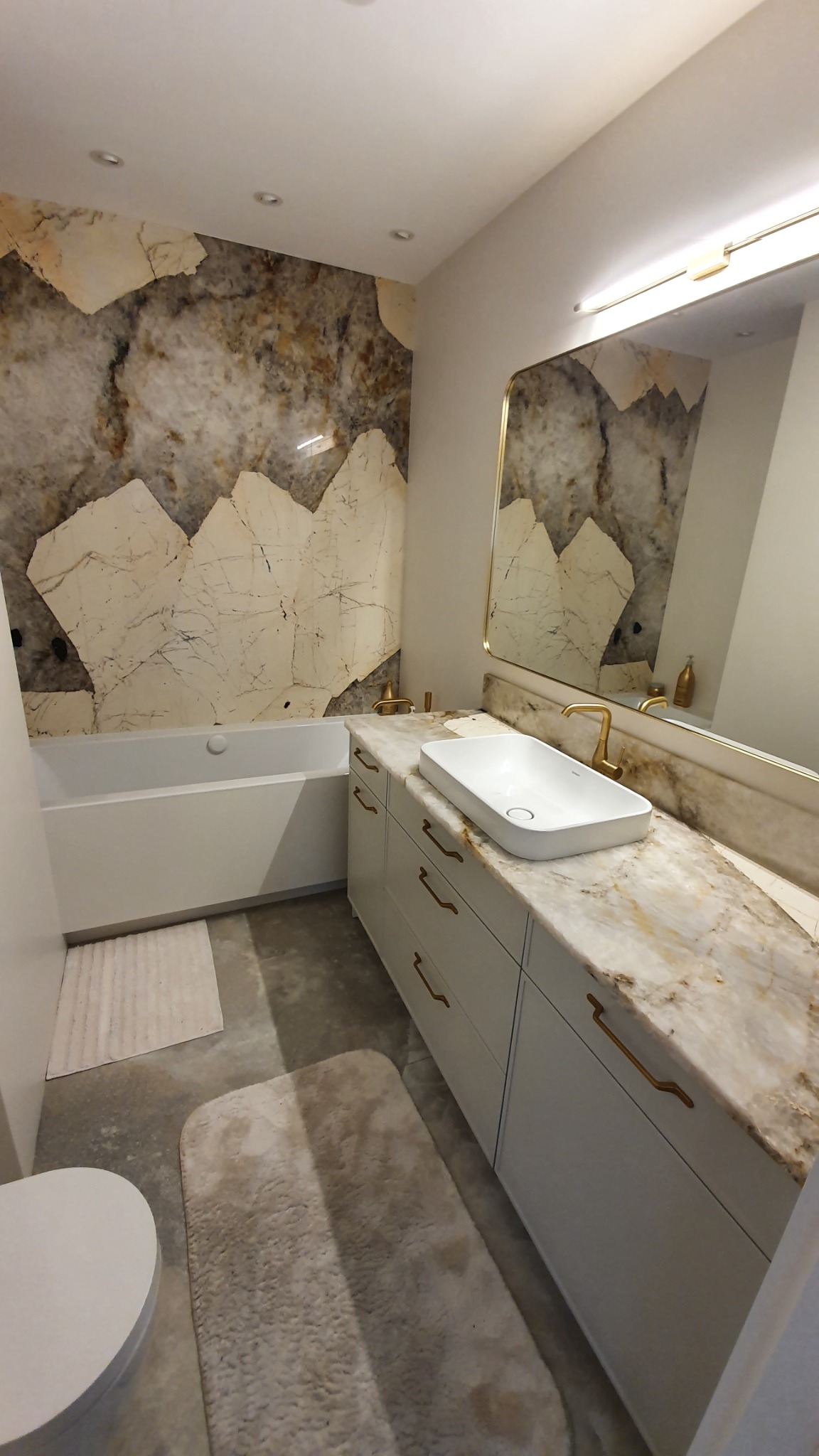 Nowoczesna łazienka z marmurową ścianą akcentującą, białą wanną i szafką z umywalką nablatową oraz złotą armaturą, oświetlona punktowo.