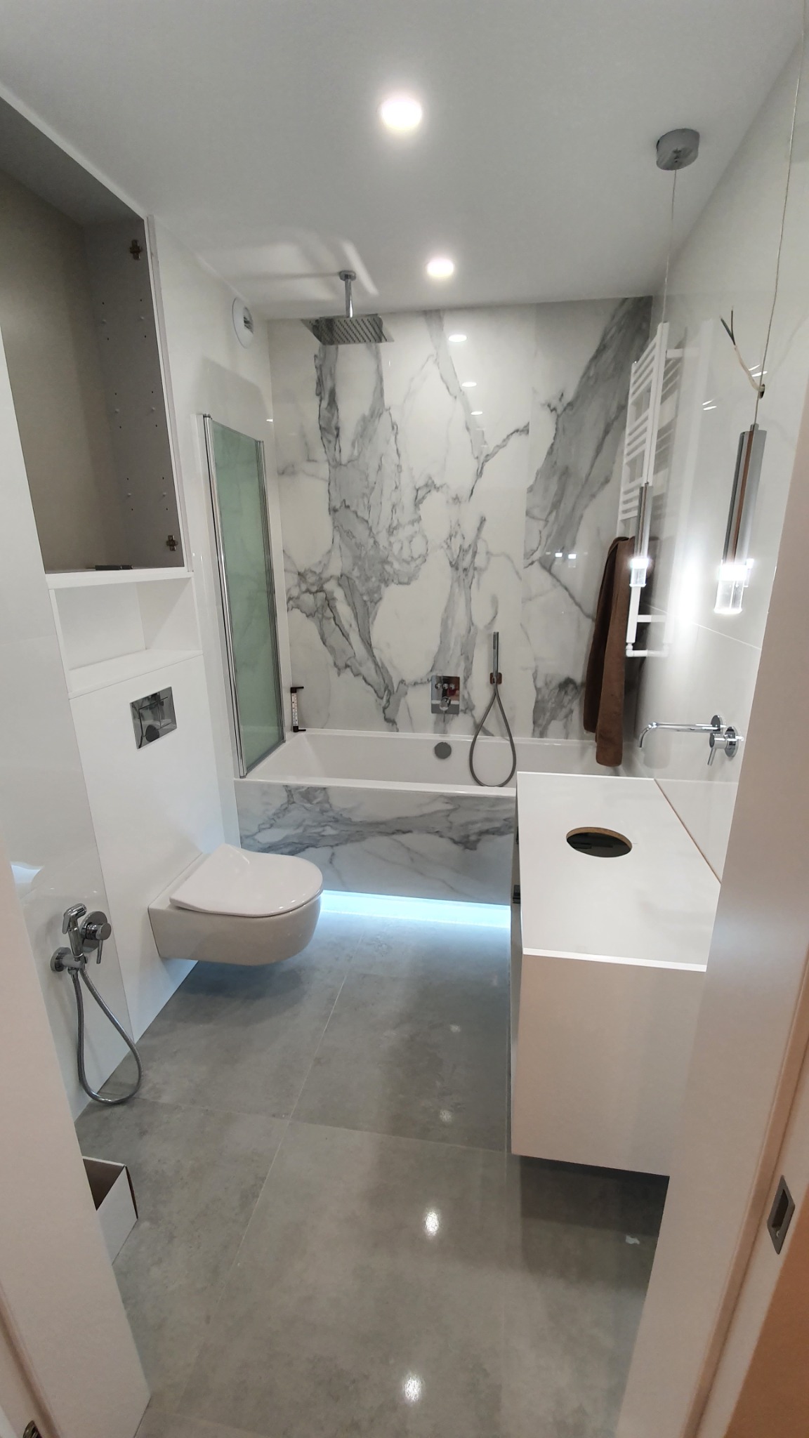 Nowoczesna łazienka z marmurową ścianą, podświetlaną wanną, podwieszaną toaletą i minimalistyczną umywalką, wykończona w bieli i szarości.