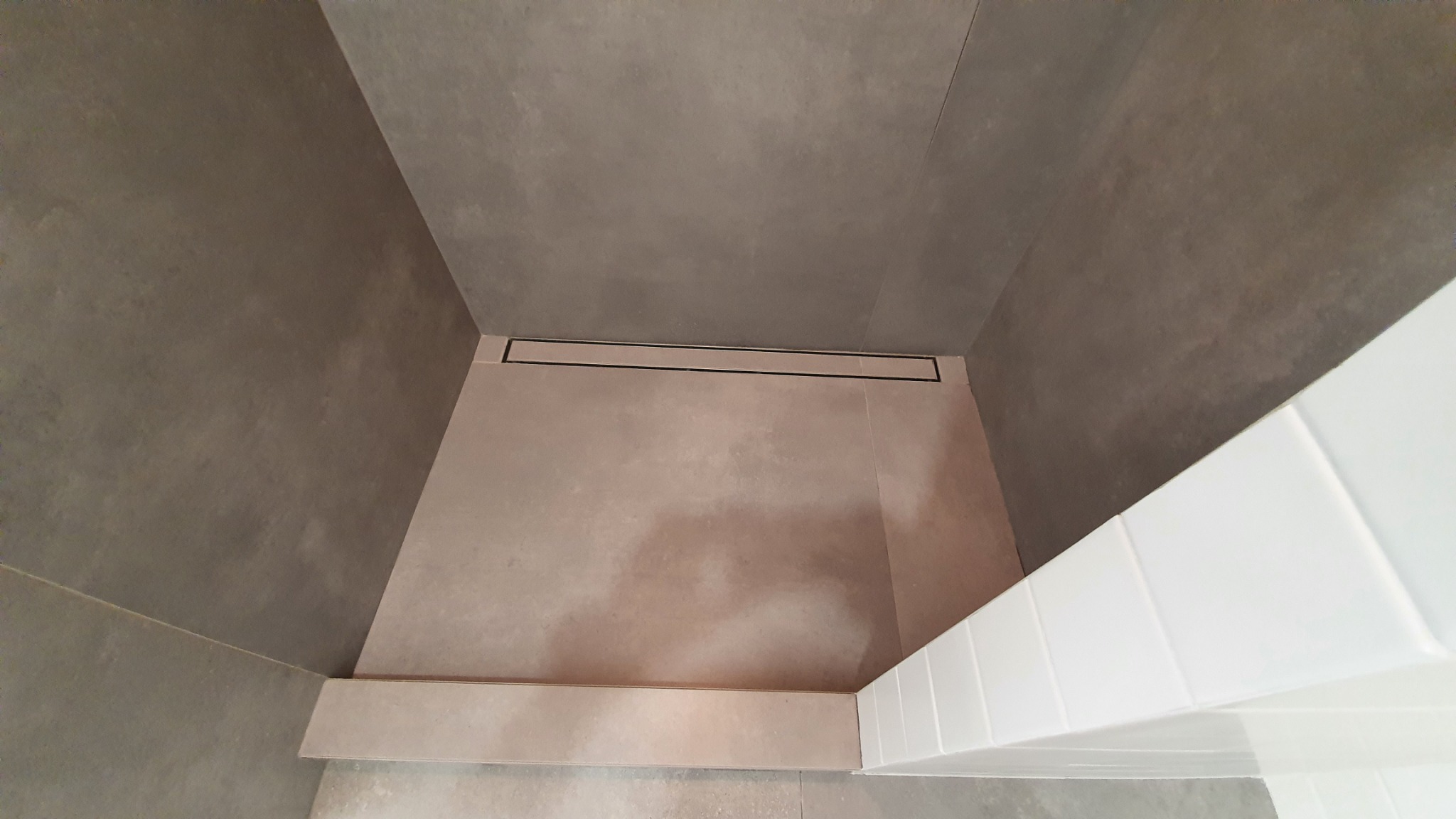 Nowoczesny brodzik wyłożony beżowymi płytkami z liniowym odpływem, otoczony szarymi płytkami ściennymi i białym obramowaniem z płytek.