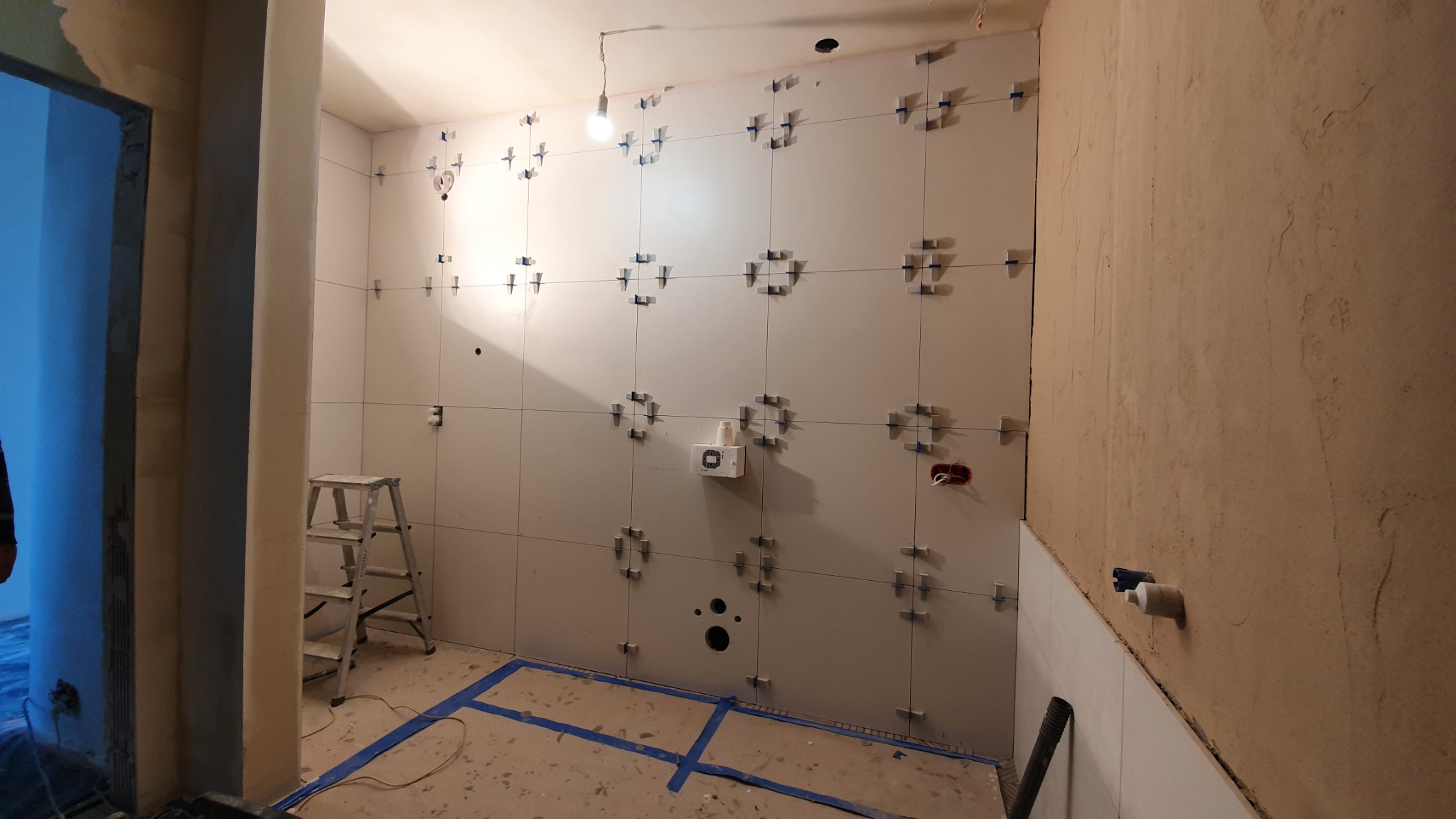 Ściany łazienki w trakcie układania płytek, widoczne krzyżyki dystansowe, drabina aluminiowa i surowa ściana obok.