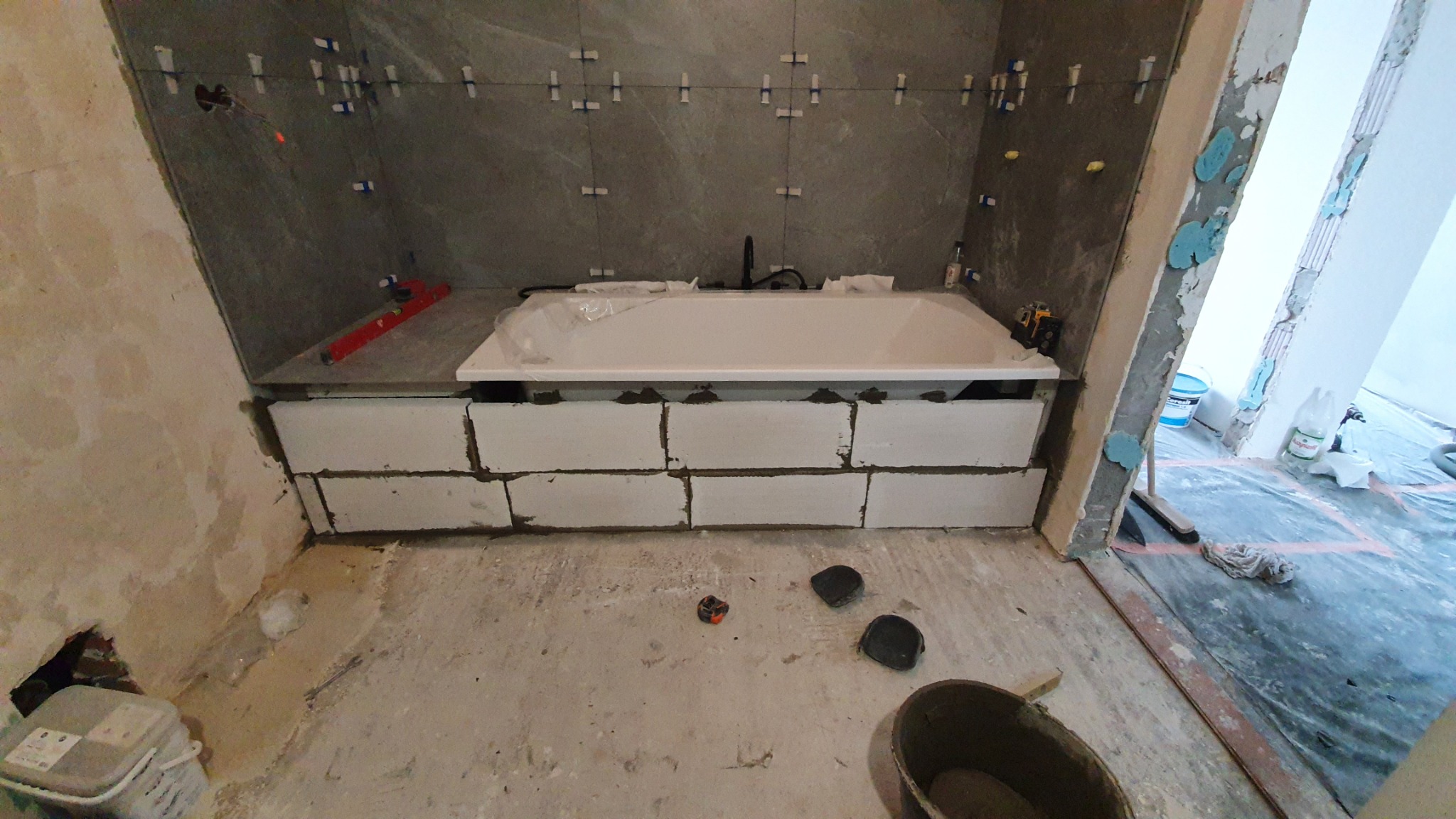 Biała wanna zabezpieczona folią, obudowana bloczkami, szare płytki na ścianie, poziomica, widoczne ślady prac remontowych, wiadro i narzędzia na podłodze.