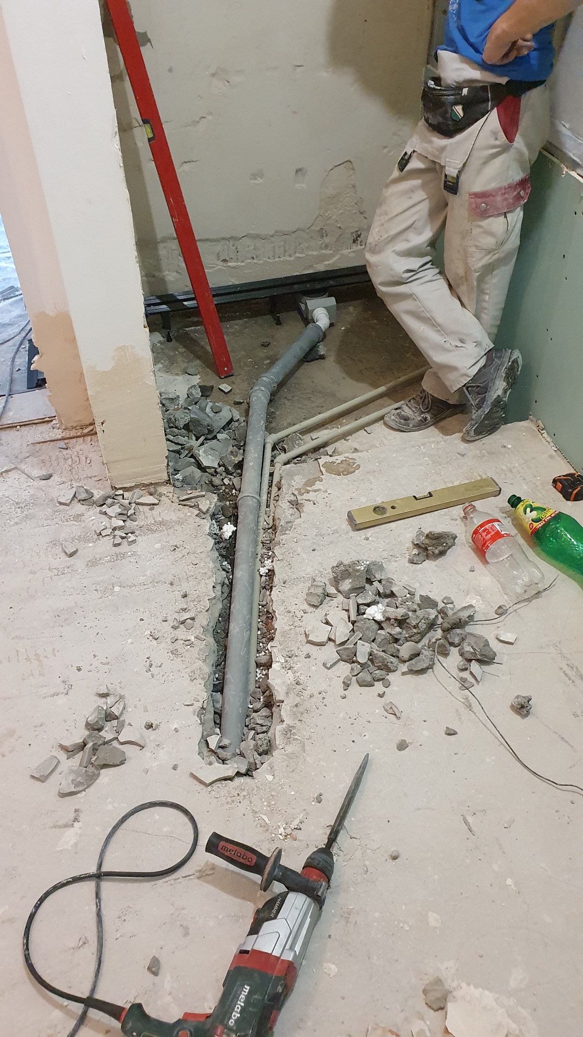 Wykuta bruzda w podłodze z szarą rurą kanalizacyjną, gruz, poziomica, perforator Metabo, instalator w roboczych spodniach oparty o ścianę.