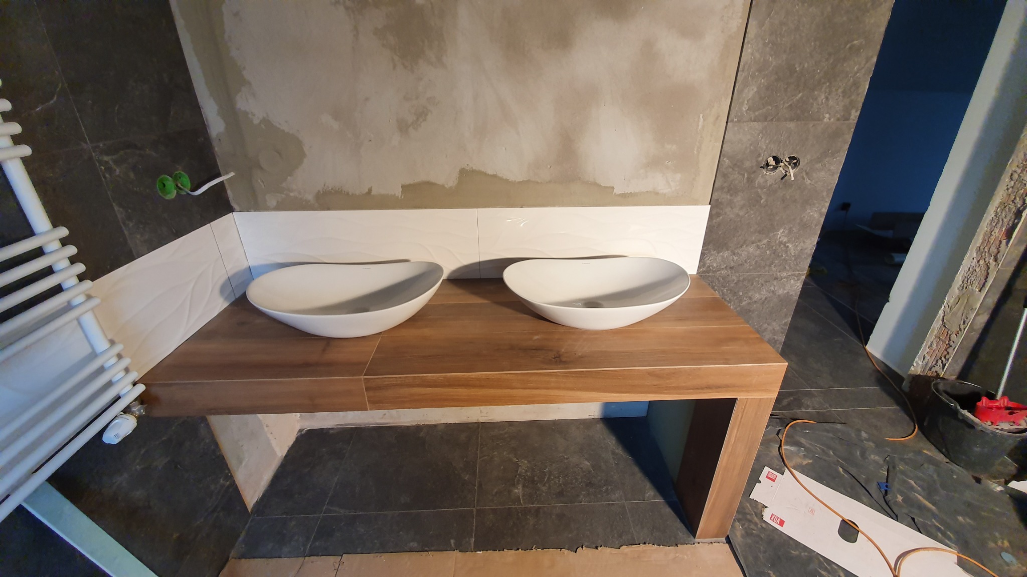 Dwie białe, owalne umywalki nablatowe, zamontowane na drewnianym blacie w łazience w trakcie remontu; widoczne płytki ścienne, grzejnik drabinkowy i niedokończone ściany.