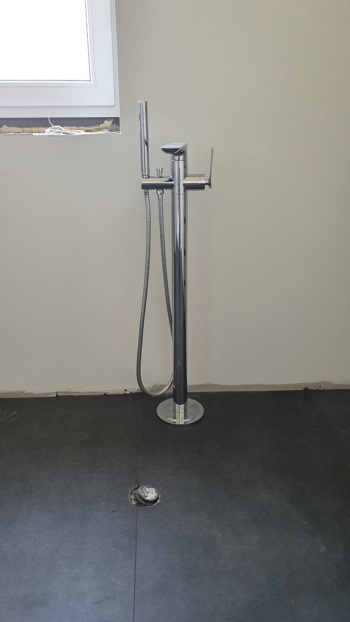 Bateria wannowa wolnostojąca chromowana, z wężem natryskowym, zamontowana na ciemnej podłodze w surowym stanie, widoczny odpływ podłogowy i niedokończone wykończenie ścian przy oknie.