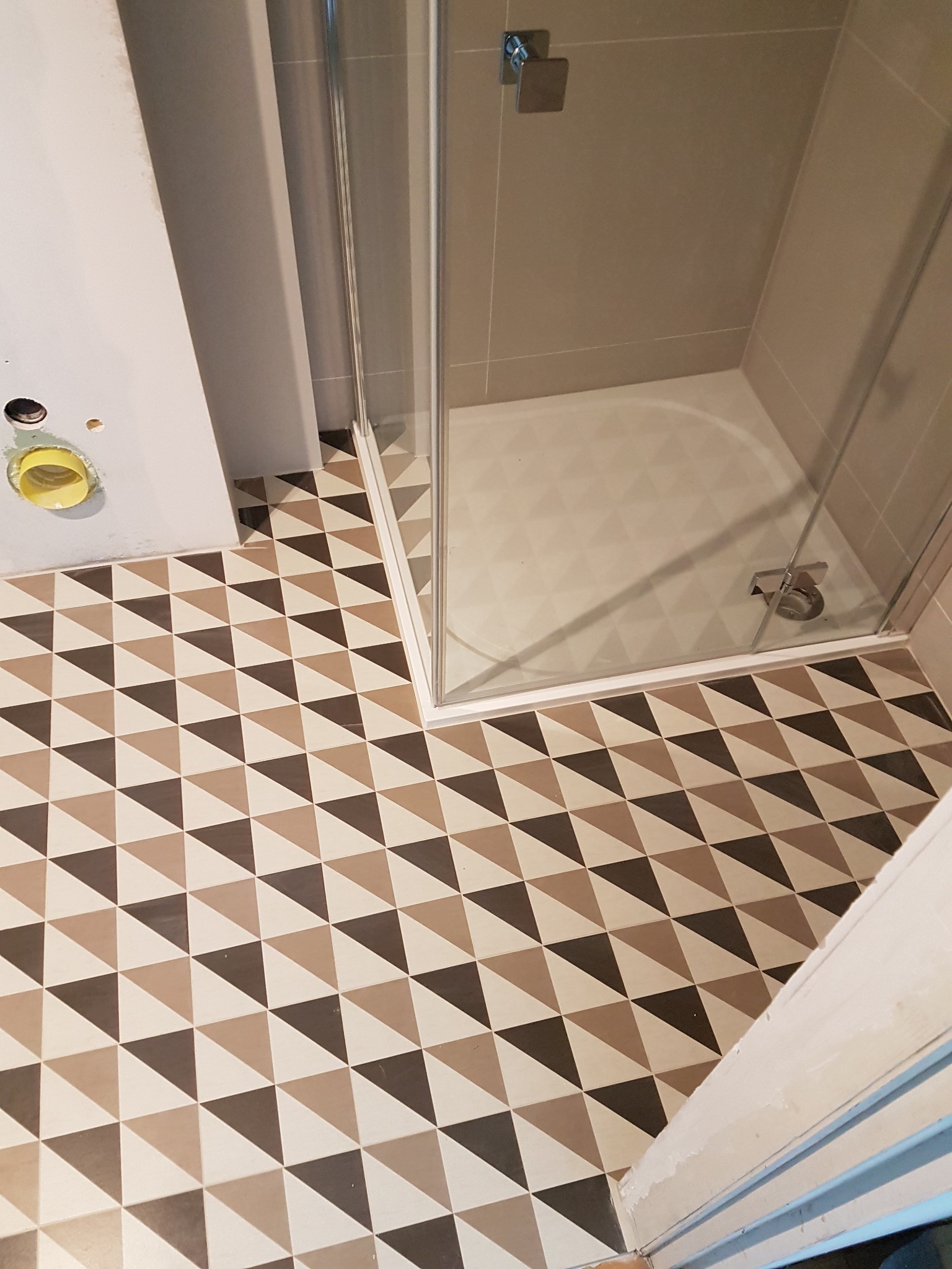 Wnętrze łazienki w trakcie remontu z mozaikową podłogą w odcieniach brązu i beżu, kabina prysznicowa z brodzikiem i widoczne przyłącze kanalizacyjne w ścianie.