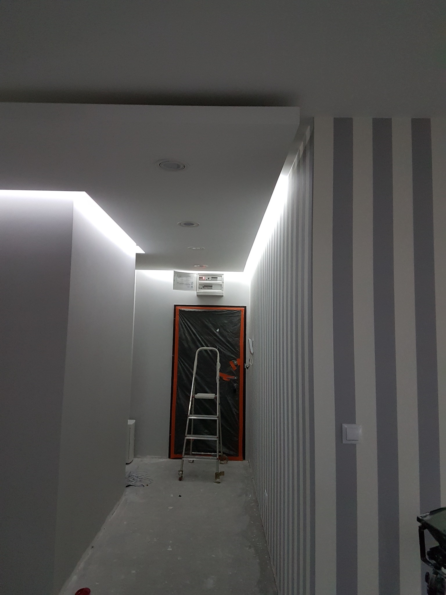 Korytarz w trakcie prac wykończeniowych: ściany pomalowane w szaro-białe pasy, sufit z oświetleniem LED, zabezpieczone drzwi folią malarską i rozstawiona drabina aluminiowa.