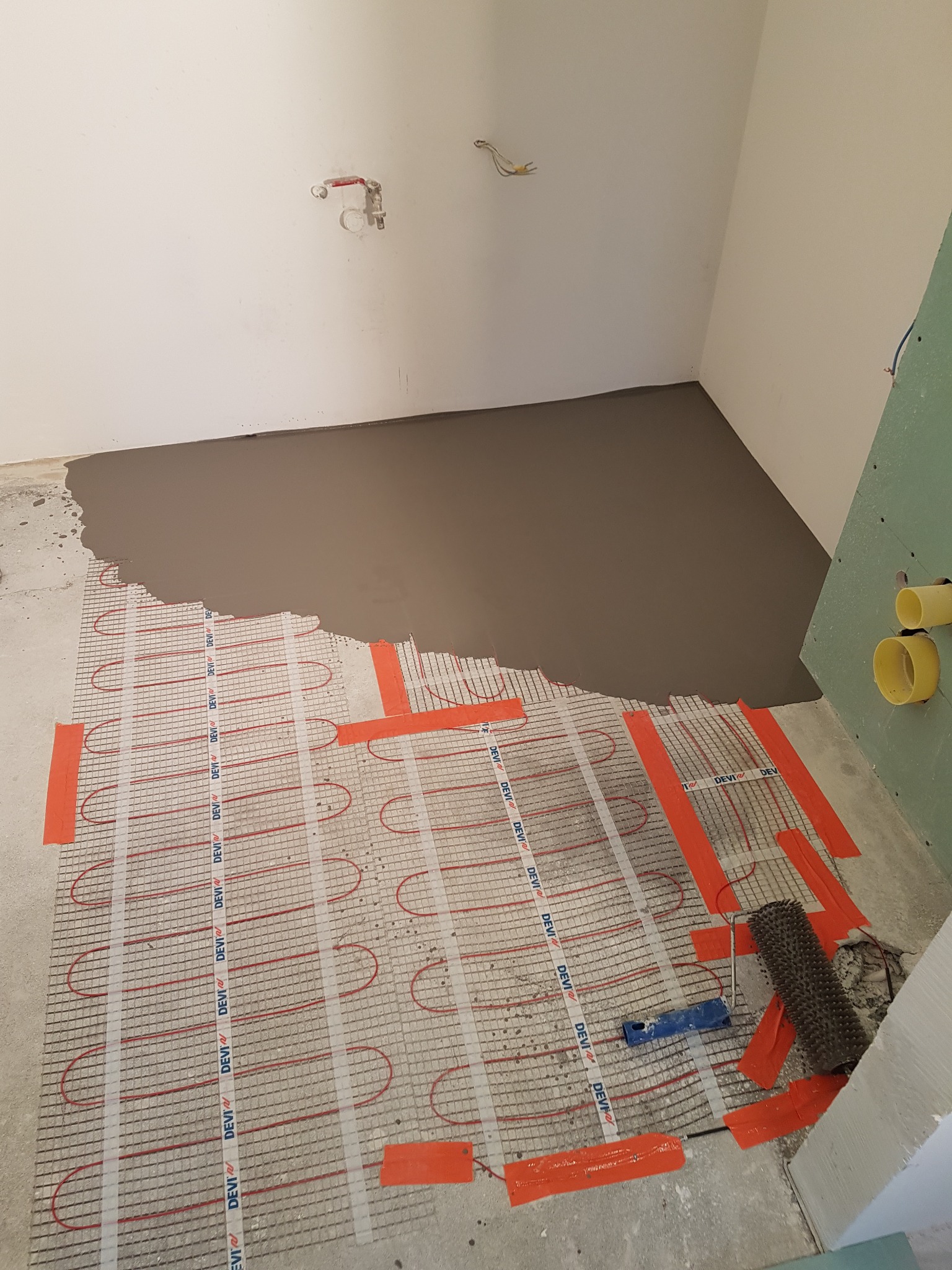 Montaż ogrzewania podłogowego: mata grzewcza DEVI z elementami mocującymi, częściowo zalana szarą wylewką samopoziomującą w surowym wnętrzu łazienki z instalacją hydrauliczną i elektryczną.