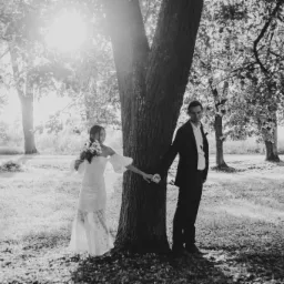 Czarno-białe zdjęcie pary młodej trzymającej się za ręce przy pniu drzewa, z promieniami słońca przebijającymi się przez liście, tworzącymi aureolę wokół drzewa.
