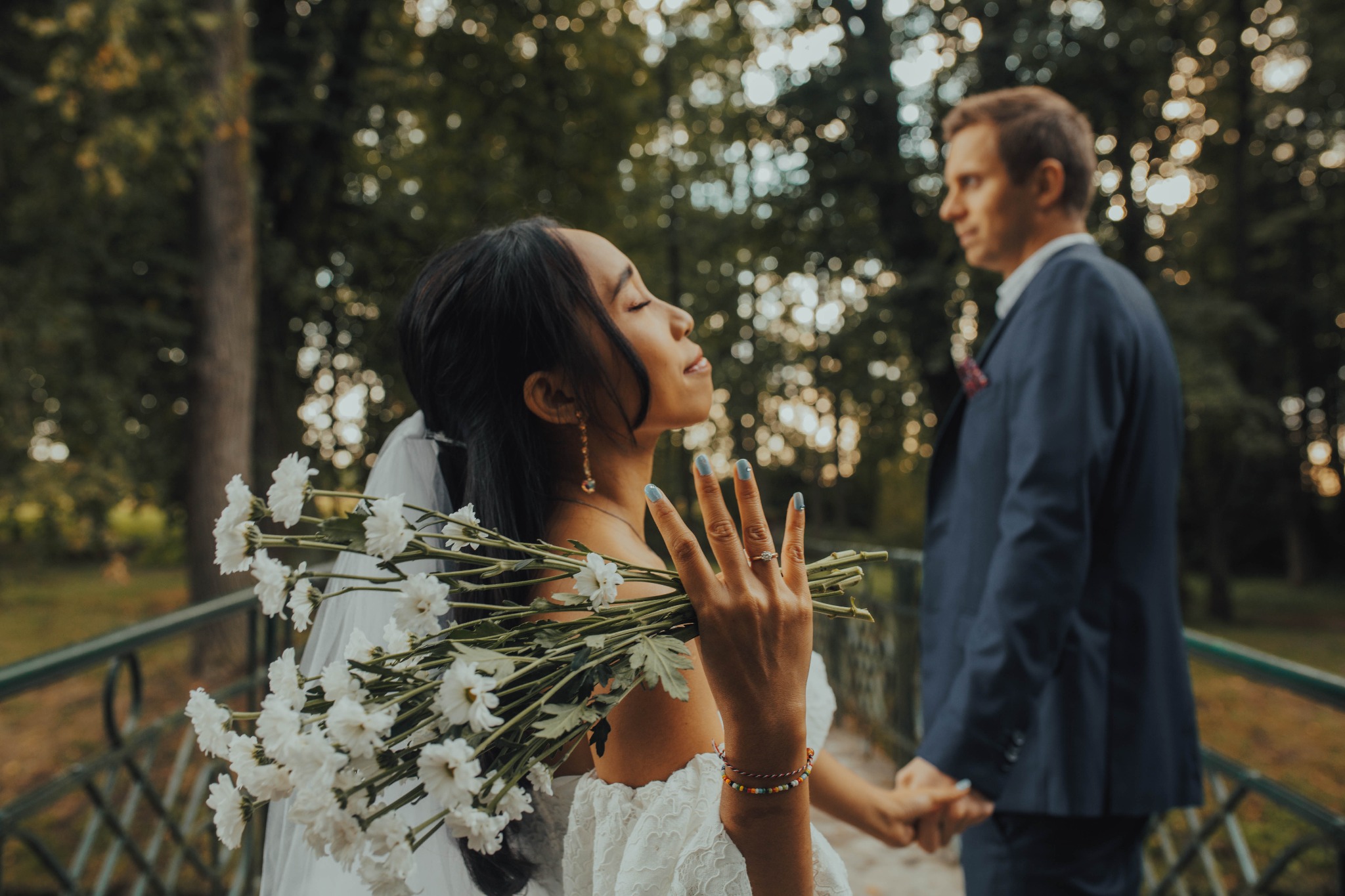 Panna młoda z zamkniętymi oczami trzyma bukiet białych chryzantem, na palcu pierścionek zaręczynowy, w tle lekko rozmyty pan młody w granatowym garniturze, oboje stoją na moście w parku.
