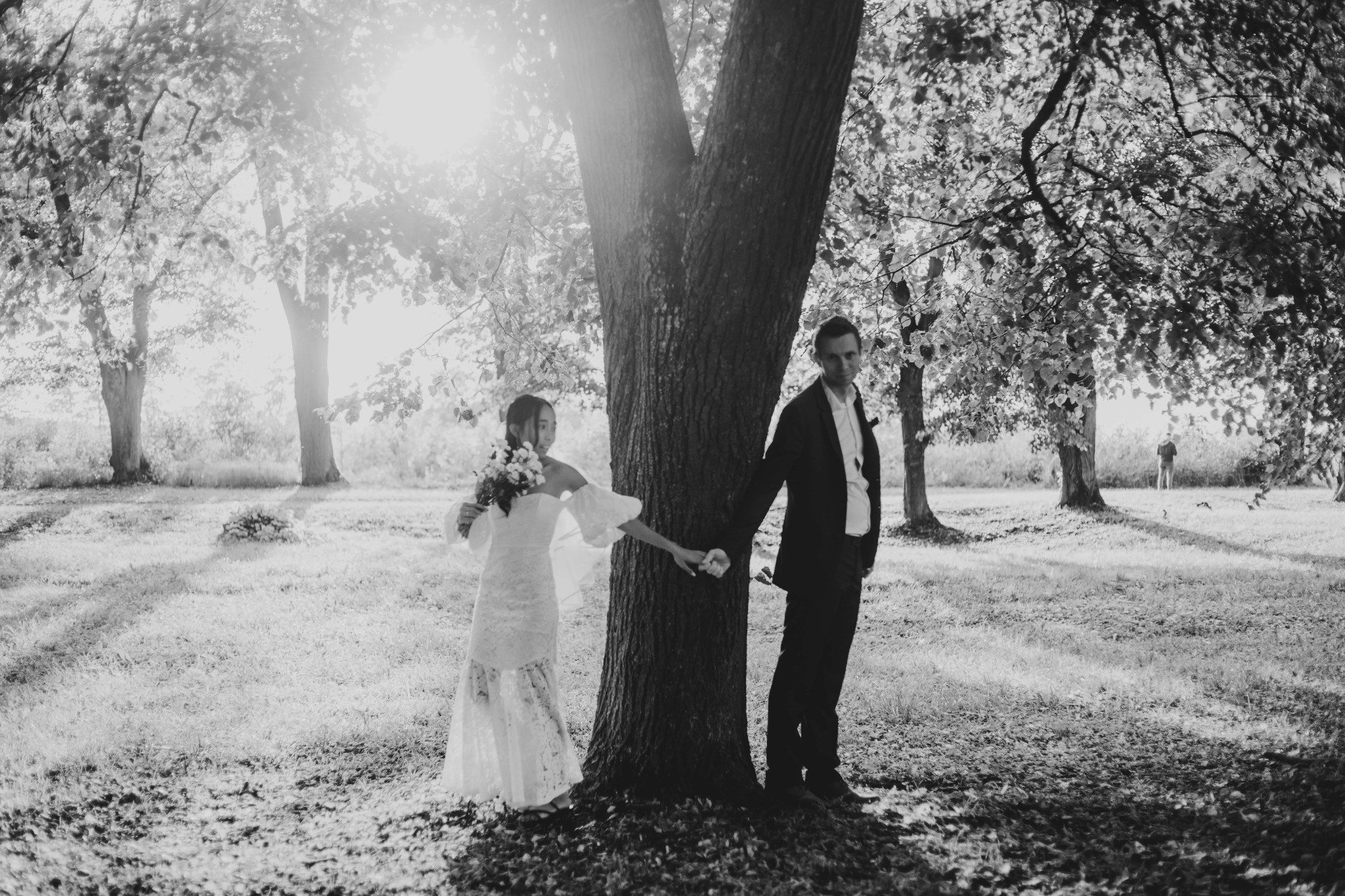 Czarno-białe zdjęcie pary młodej trzymającej się za ręce przy pniu drzewa, z promieniami słońca przebijającymi się przez liście, tworzącymi aureolę wokół drzewa.