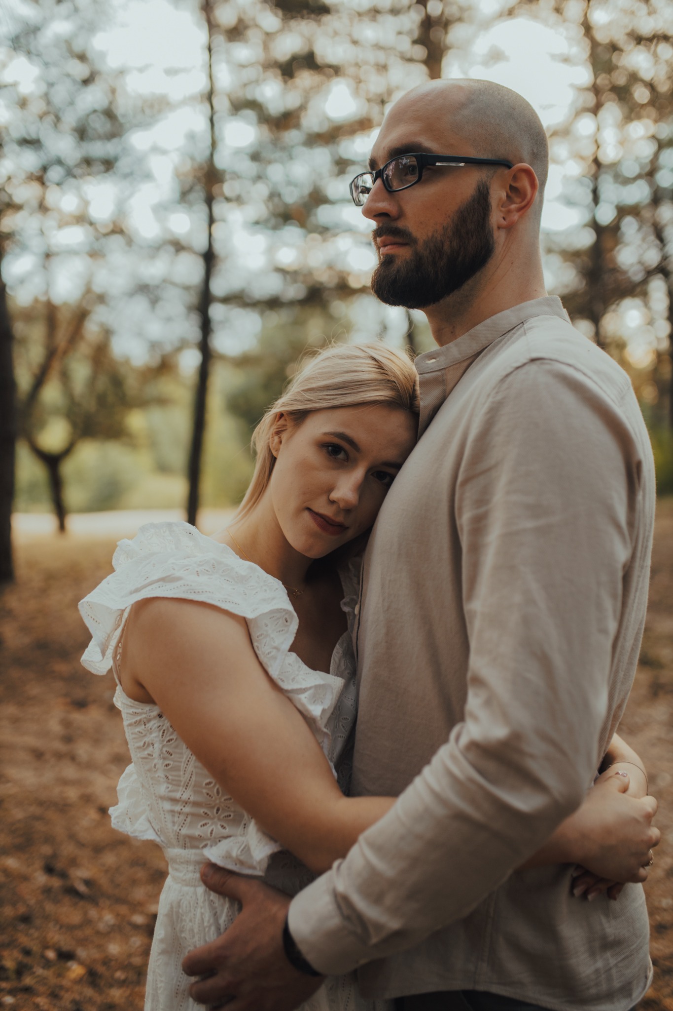 Para zakochanych w objęciach na tle leśnej scenerii: ona w białej, ażurowej sukience, on w beżowej koszuli, z brodą i okularami.