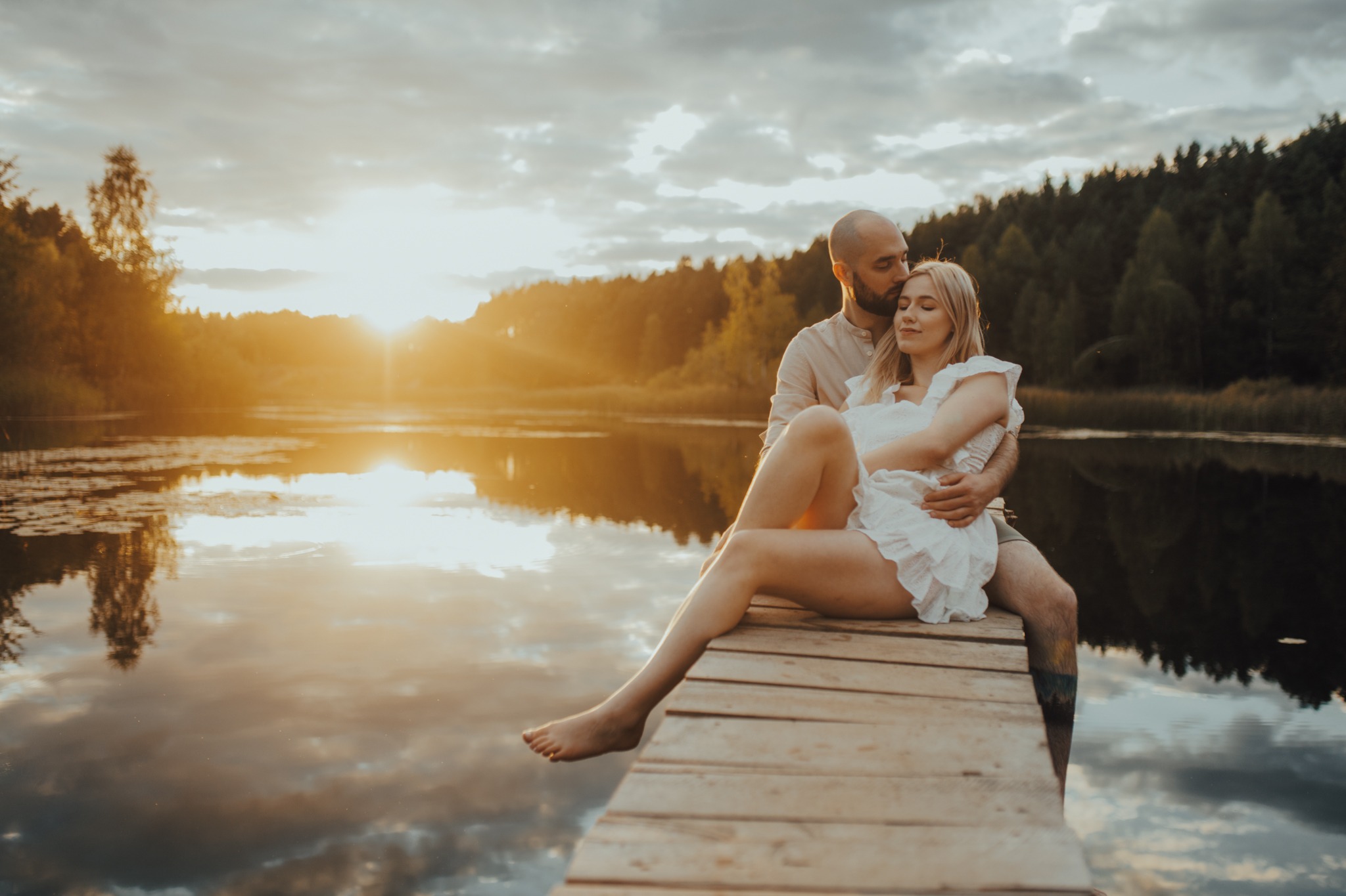 Para siedząca na drewnianym pomoście nad jeziorem o zachodzie słońca, mężczyzna obejmuje kobietę ubraną w białą sukienkę z falbanami.